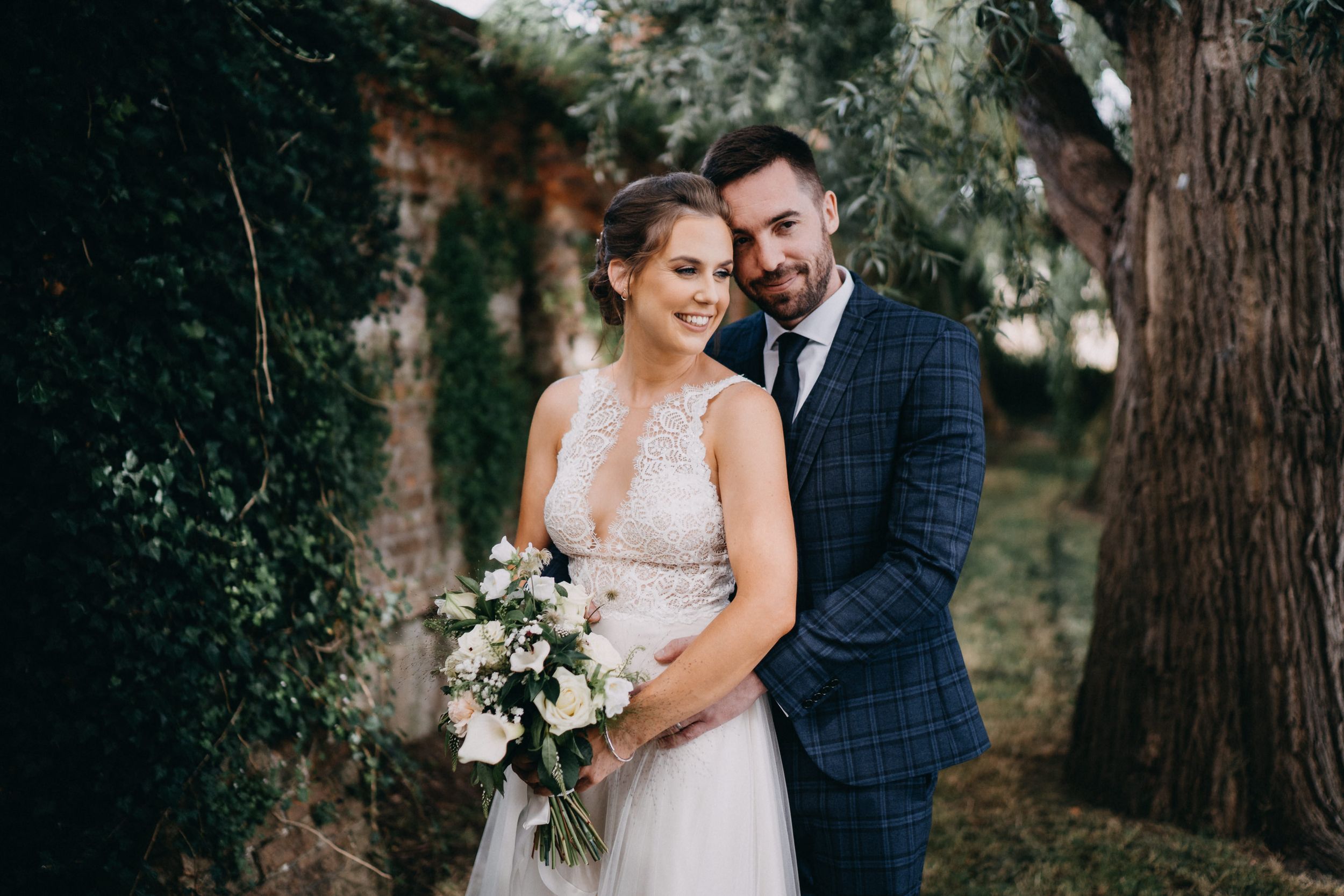 A bride and groom stand together outdoors holding a white floral bouquet against a natural woodland backdrop.