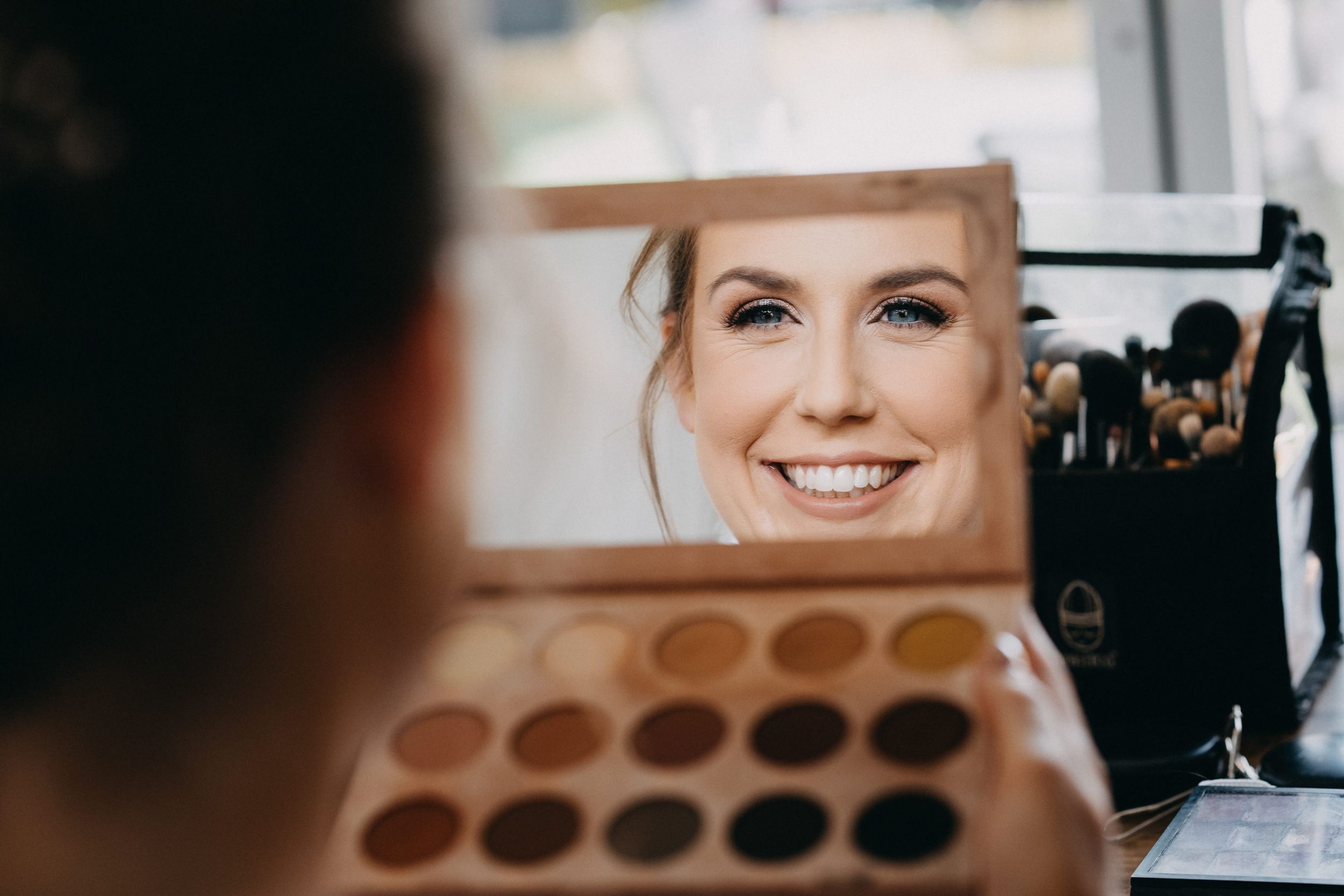 A reflection in a mirror shows a bright smile while an eyeshadow palette is visible in the foreground.