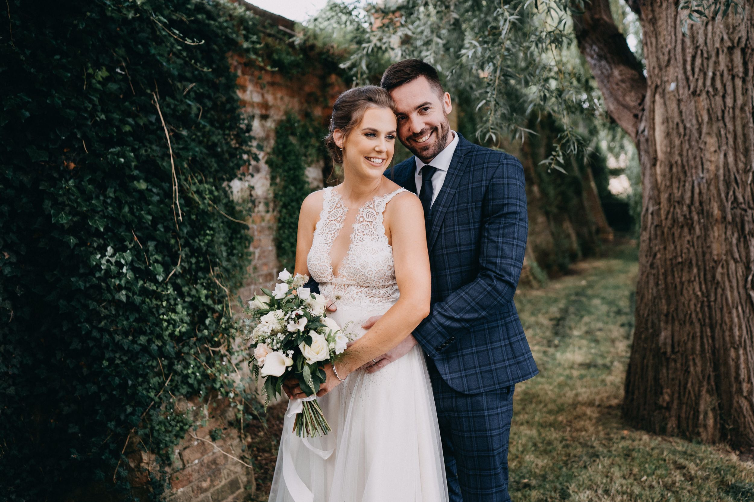 A couple in wedding attire poses together in a garden setting of The Old Bridge Hotel with trees and a white floral bouquet.