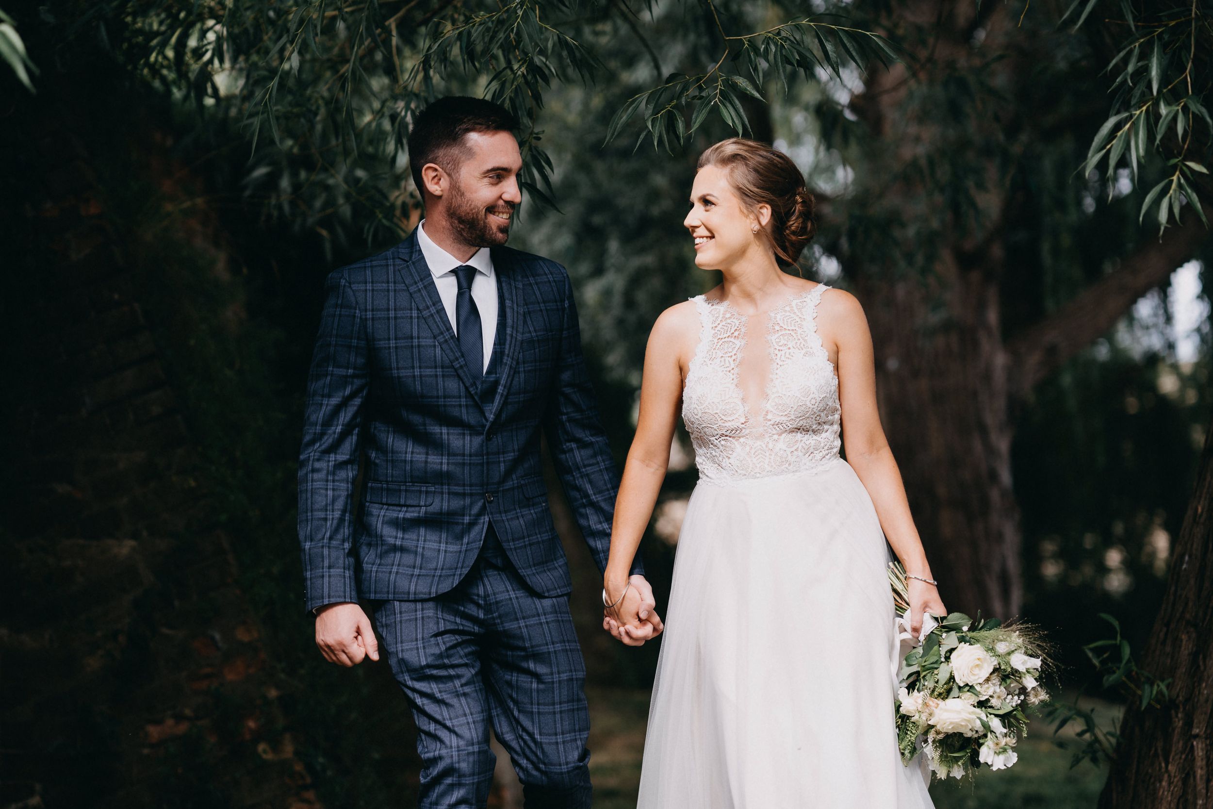 Newlywed couple holding hands and walking together outdoors, with bride in flowing white gown and groom in navy suit.
