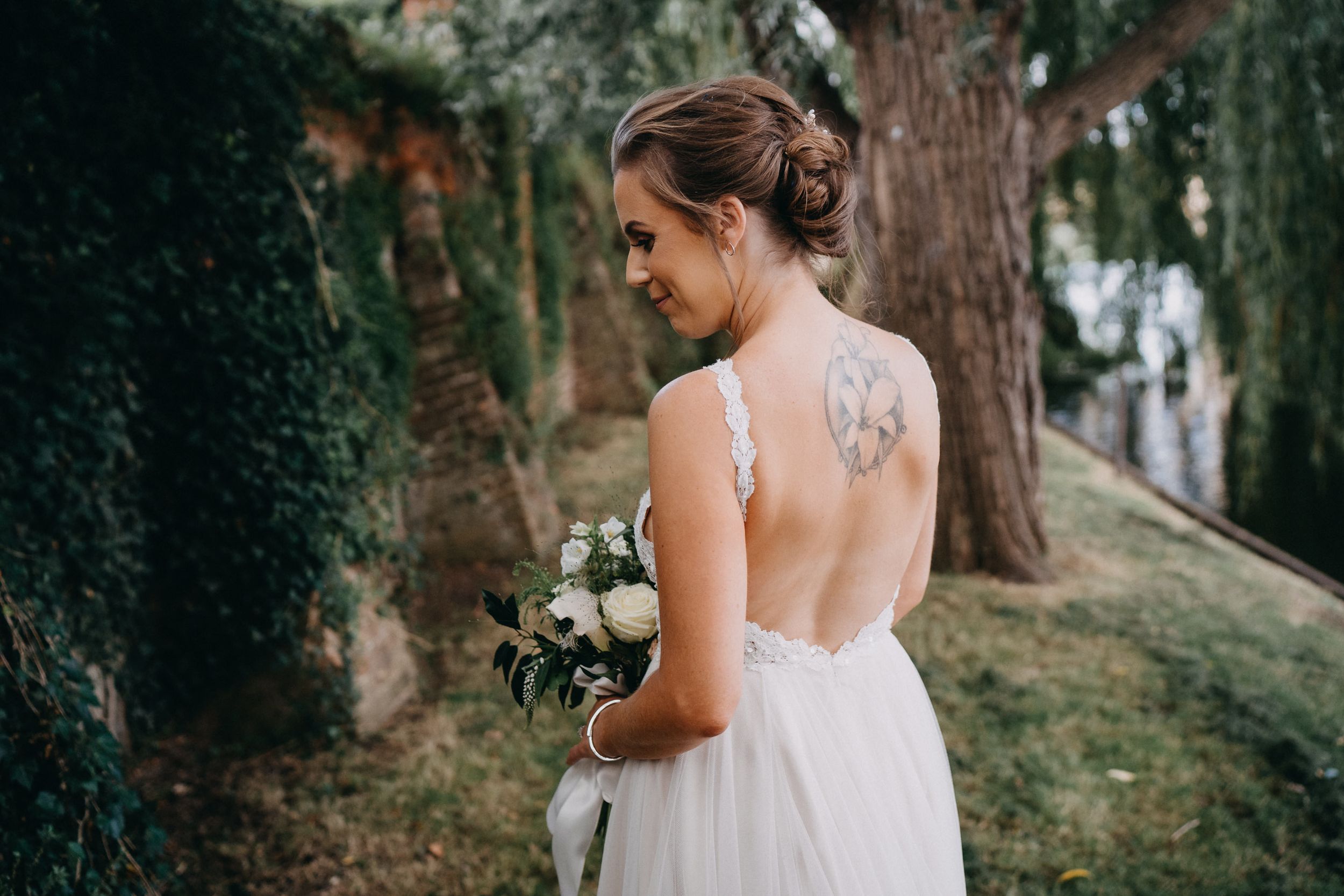 Bride in sleeveless open-back white wedding dress stands among trees holding bouquet in outdoor garden setting.
