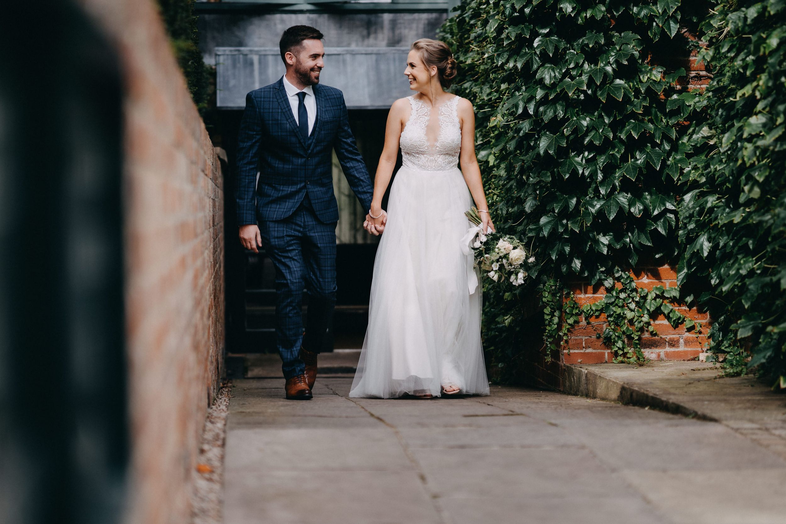 A bride and groom walk together along a stone path bordered by climbing ivy and greenery.