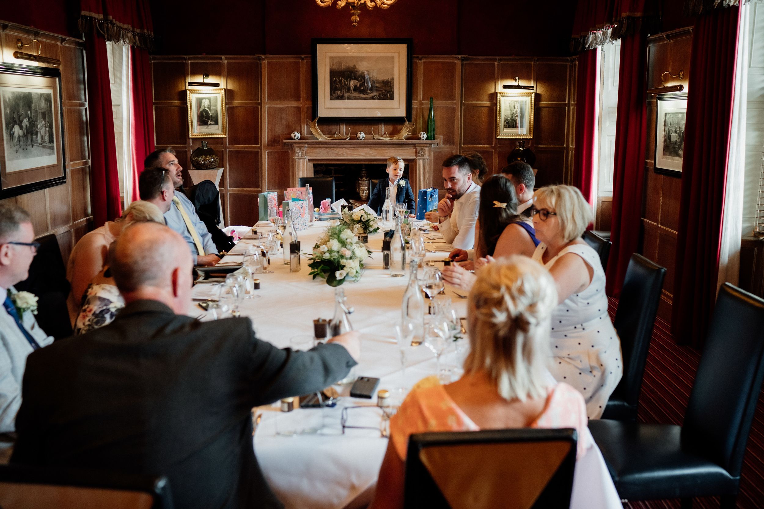 Wedding reception guests gather around an elegant table in a wood-paneled dining room with portrait art on the walls.
