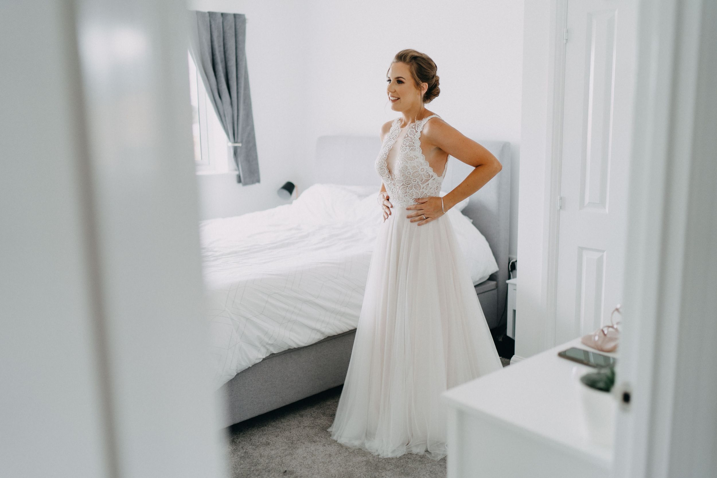 A person stands in an elegant white wedding gown in a bright bedroom, photographed through a doorway.