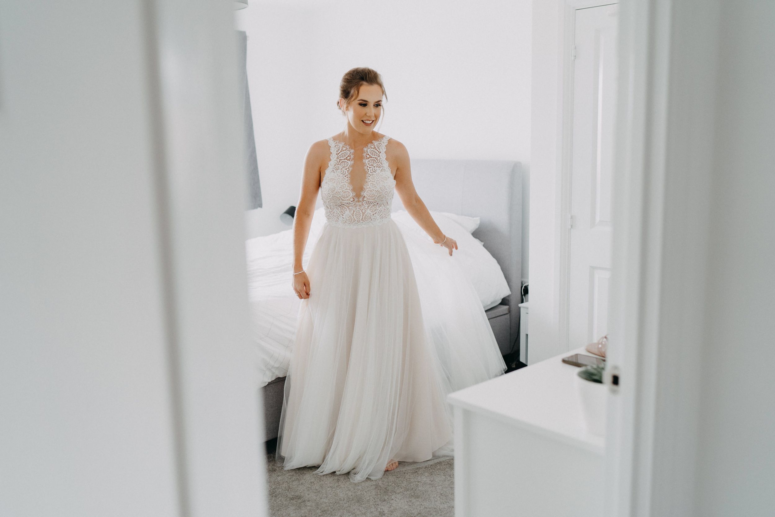 A bride wearing an elegant white flowing wedding gown with a plunging neckline and draped chiffon skirt stands in a bright room.