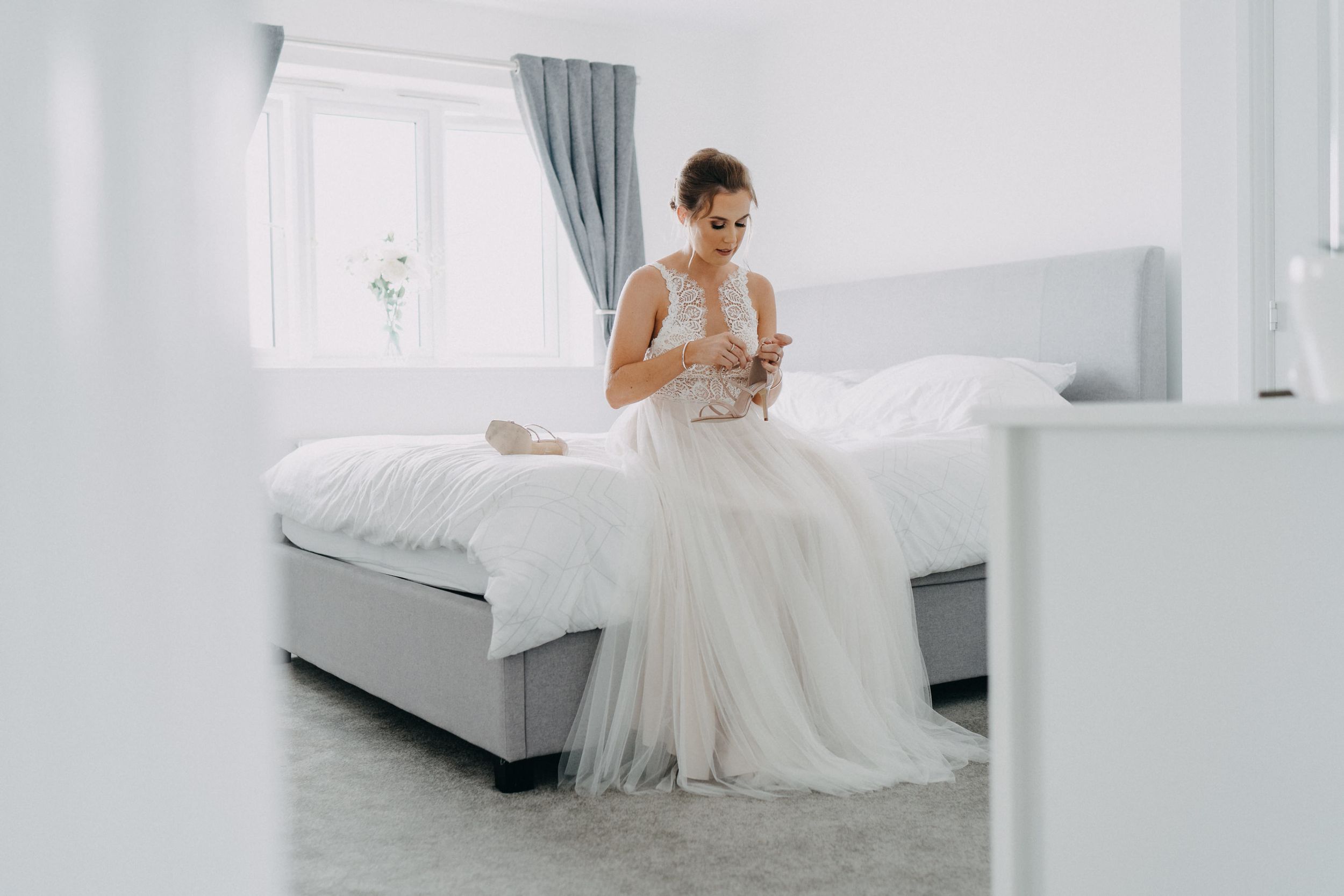 A figure in a flowing white wedding gown sits on a light gray bed in a bright, minimalist bedroom setting.