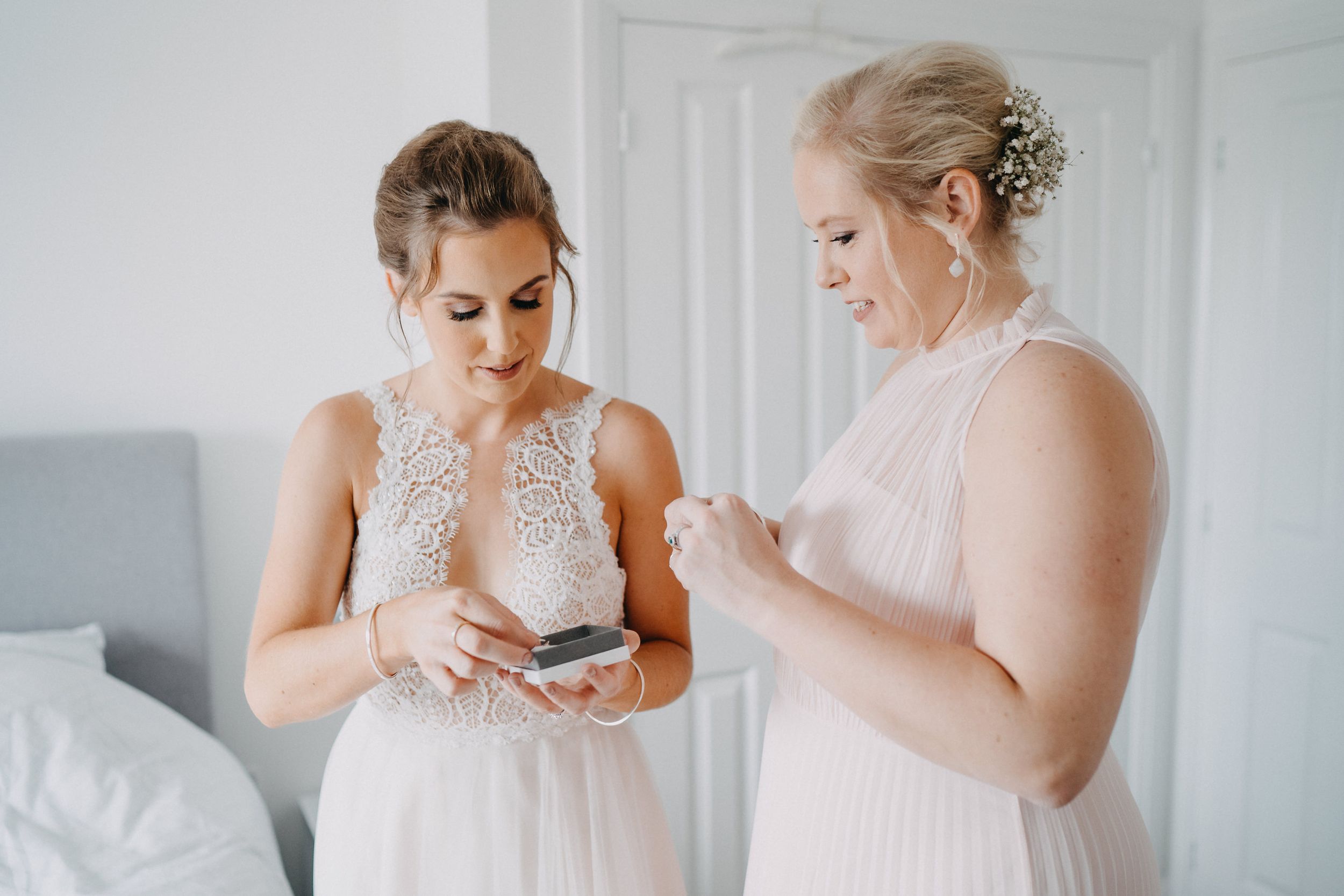 Two women in elegant light-colored dresses look at their phones while getting ready for a formal event.