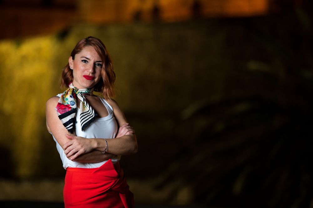 A person in a red skirt and floral top poses confidently against a dark background with dramatic lighting.