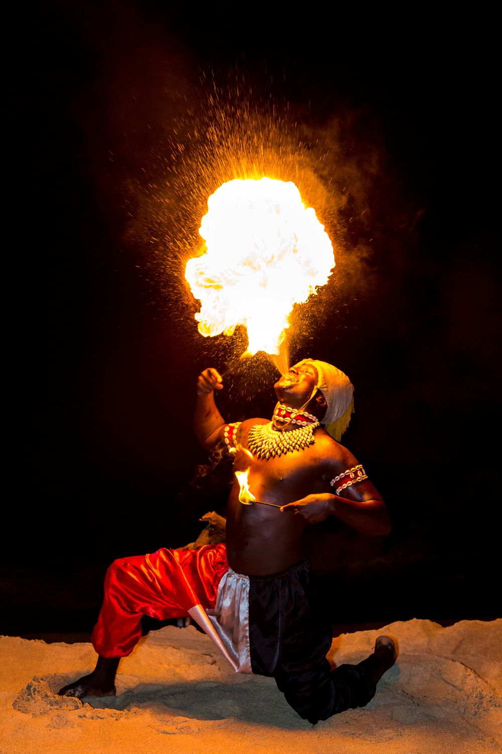 A fire performer breathes flames into the night sky while kneeling on snowy ground in red pants.