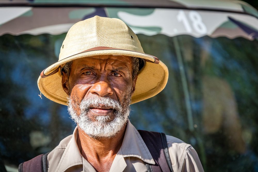 An elderly person wearing a tan safari hat and button-down shirt smiles warmly at the camera.