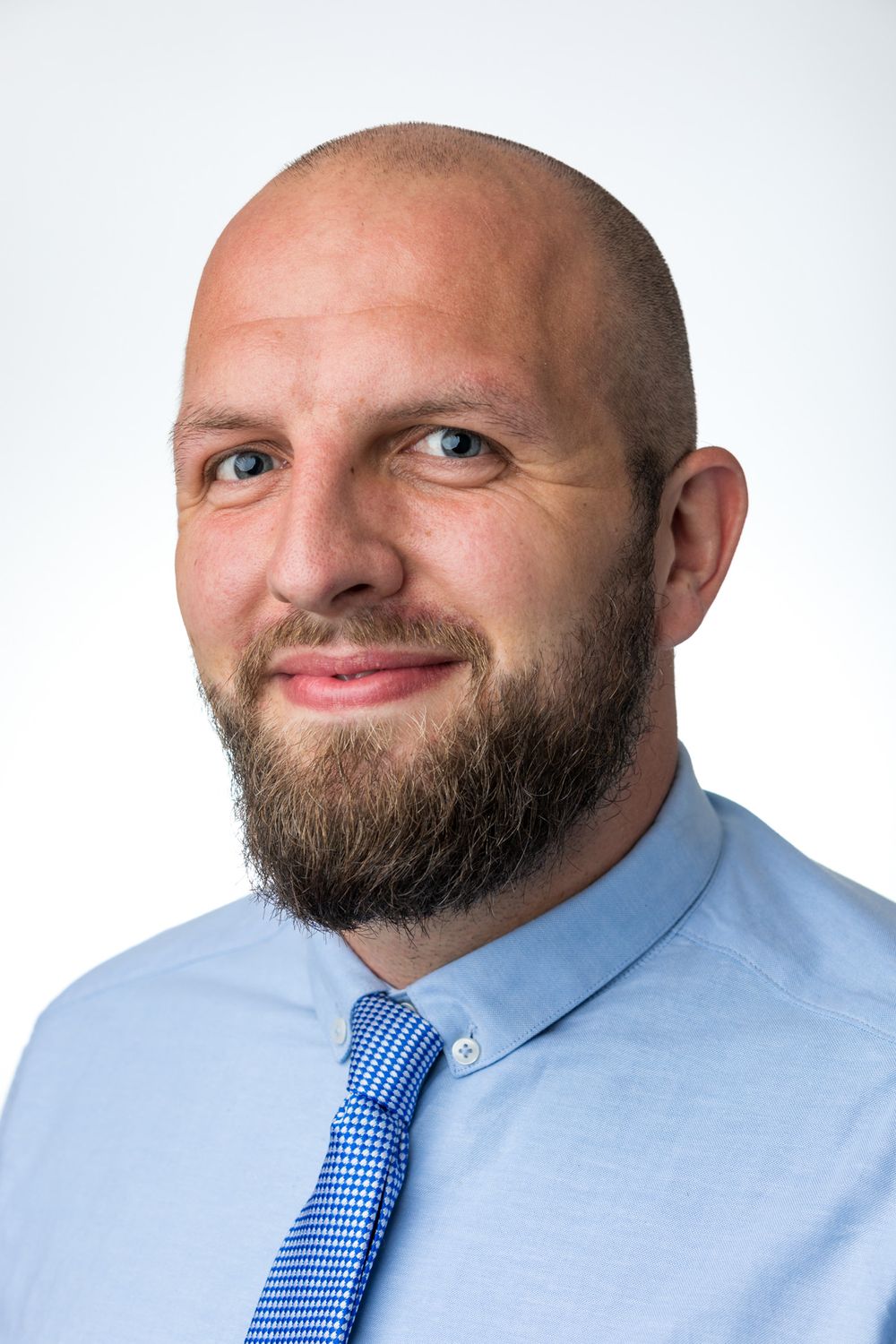 Professional headshot of bearded person in blue shirt and tie against white background.