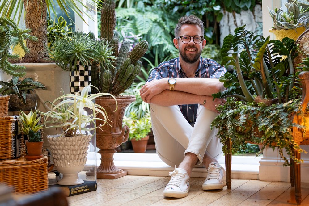 Someone sits surrounded by lush indoor plants and decorative planters in a bright sunlit interior space.