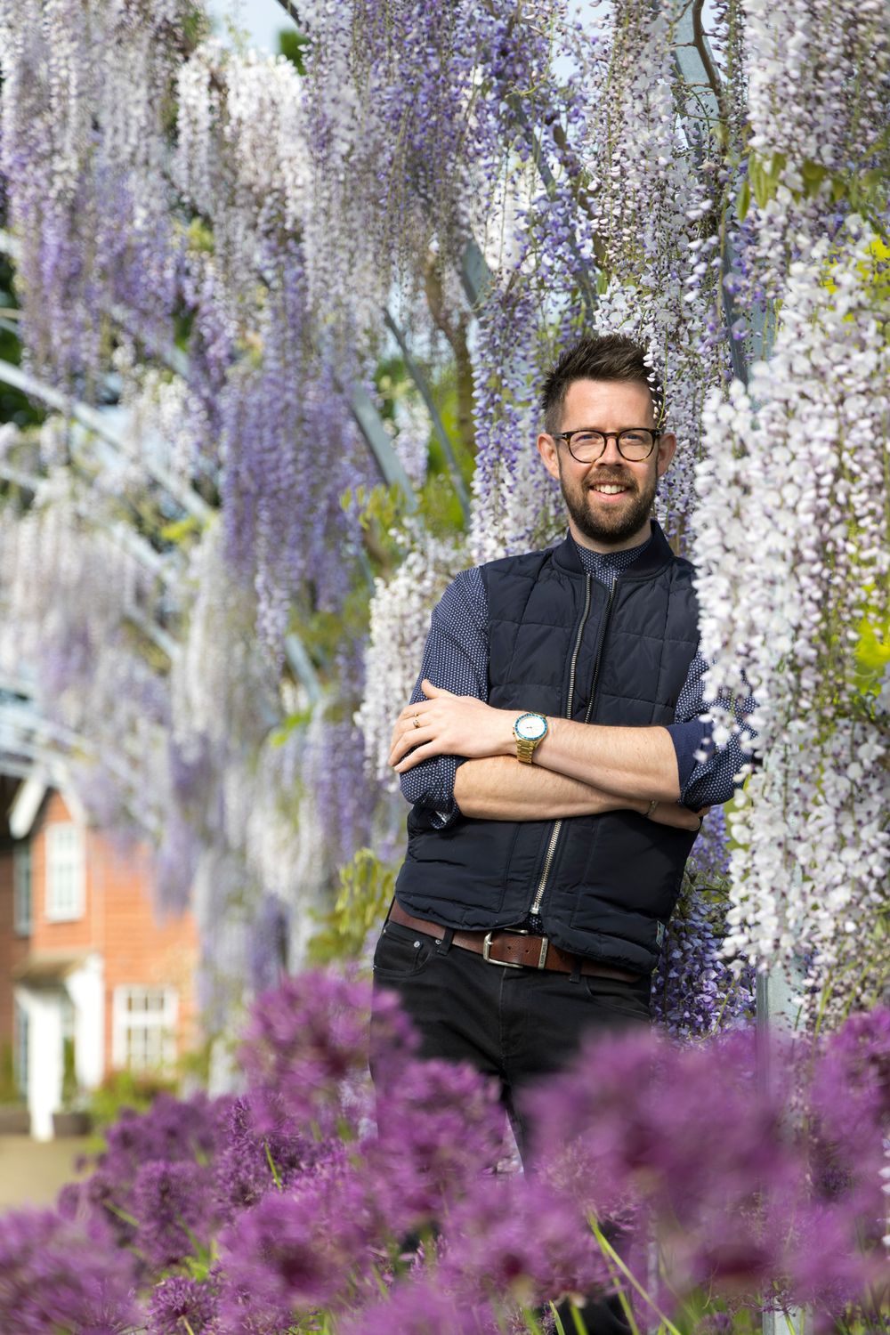 A person stands among blooming purple wisteria vines wearing a dark button-up shirt and glasses.