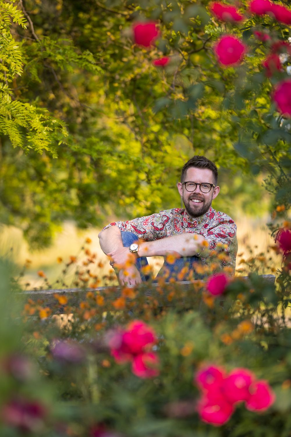 Person smiling in a garden with vibrant pink roses and lush greenery in soft natural lighting.