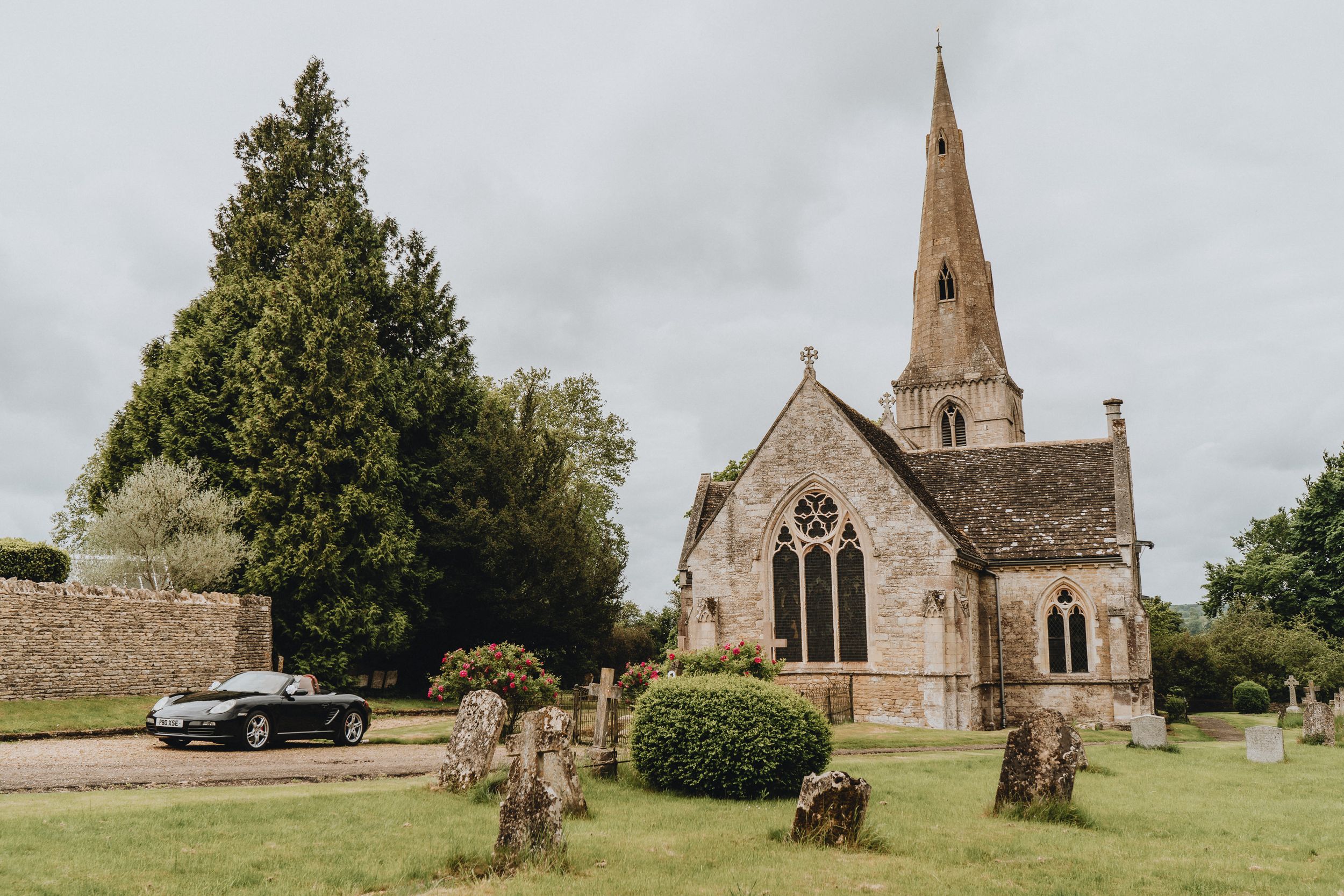 A picturesque stone church with spire stands among trees and gravestones in a rural English countryside setting.