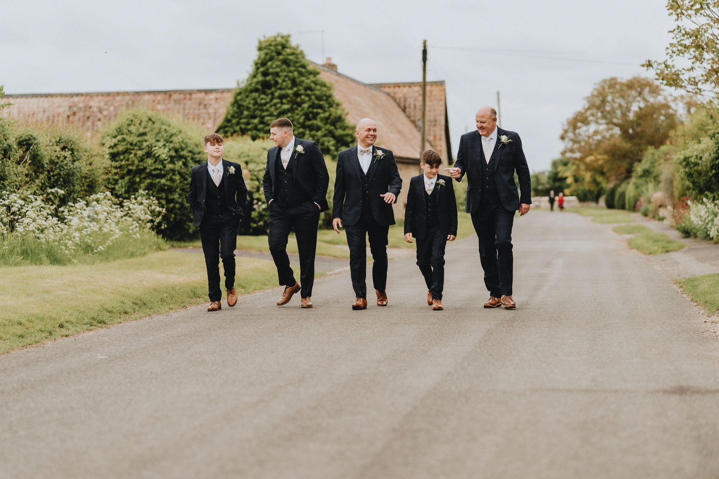 A group of five groomsmen in black suits walking together on a country road near a rustic building.