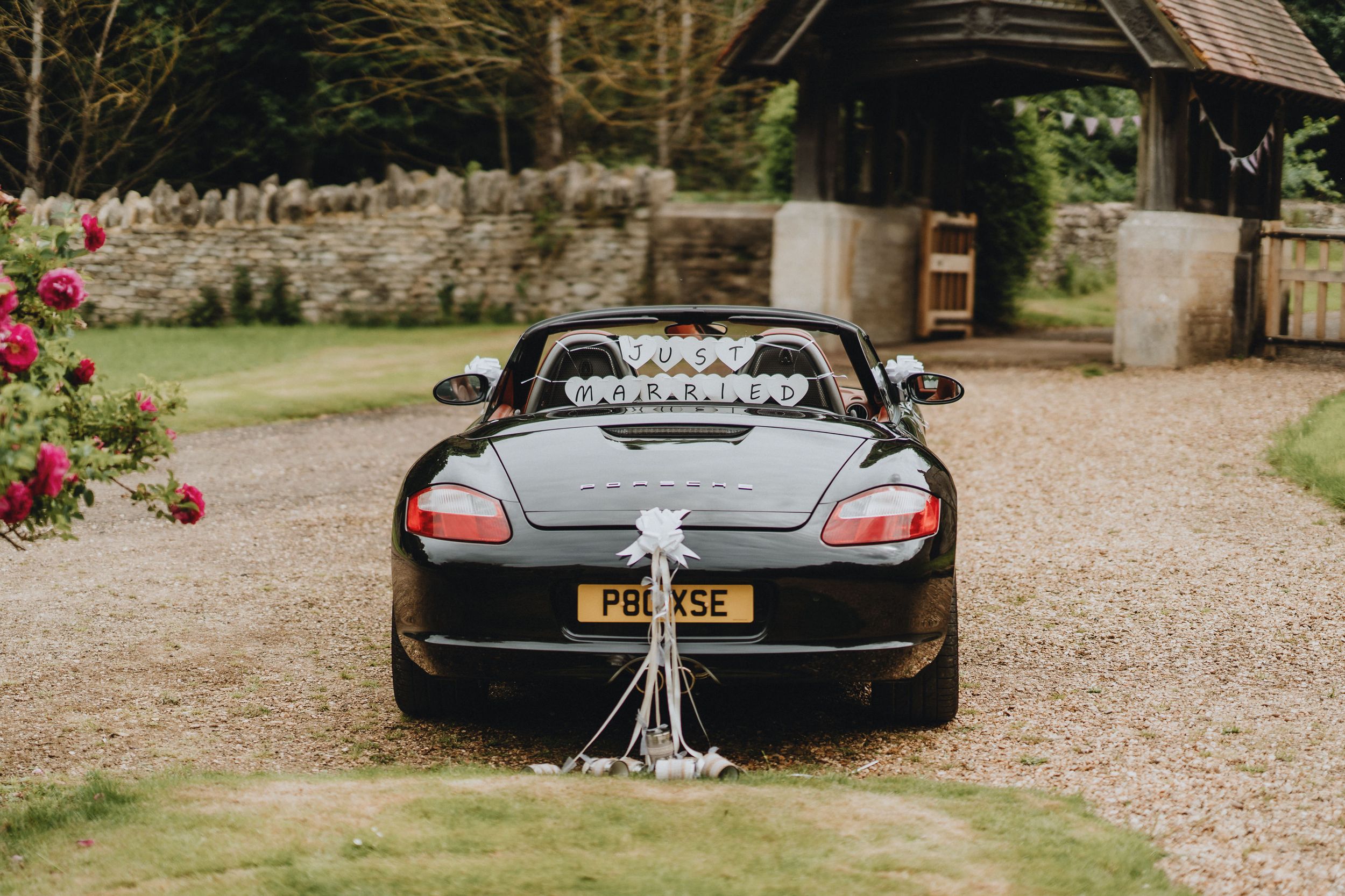 Sleek black Mercedes convertible decorated with wedding ribbons and cans parked on gravel driveway near stone wall and roses.
