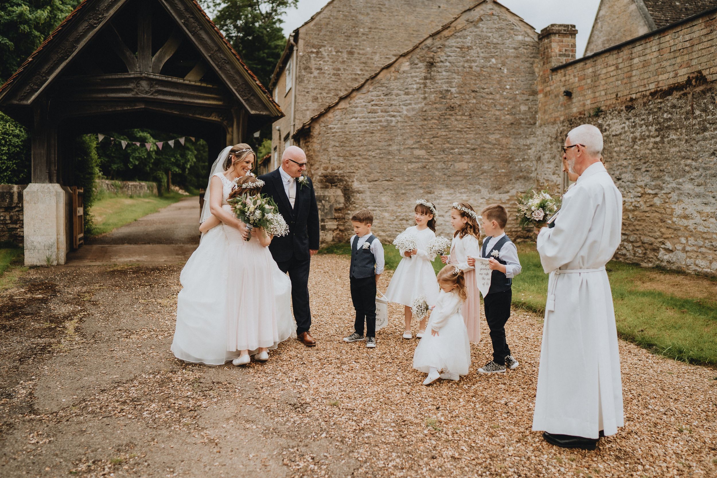 Wedding ceremony takes place under a rustic wooden lychgate with people gathered on a gravel path.