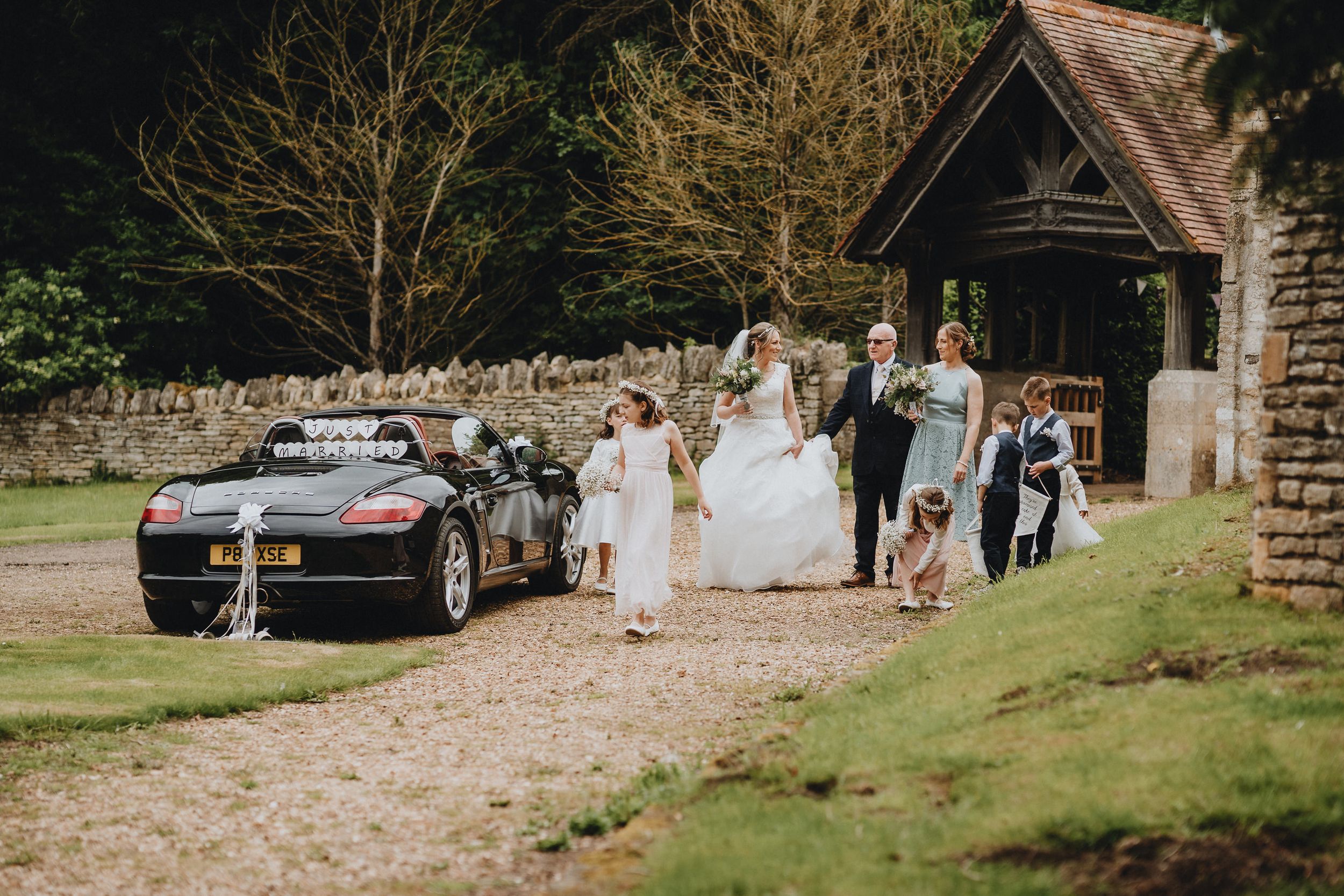 Wedding party exits stone chapel entrance with decorated black car parked on gravel path.