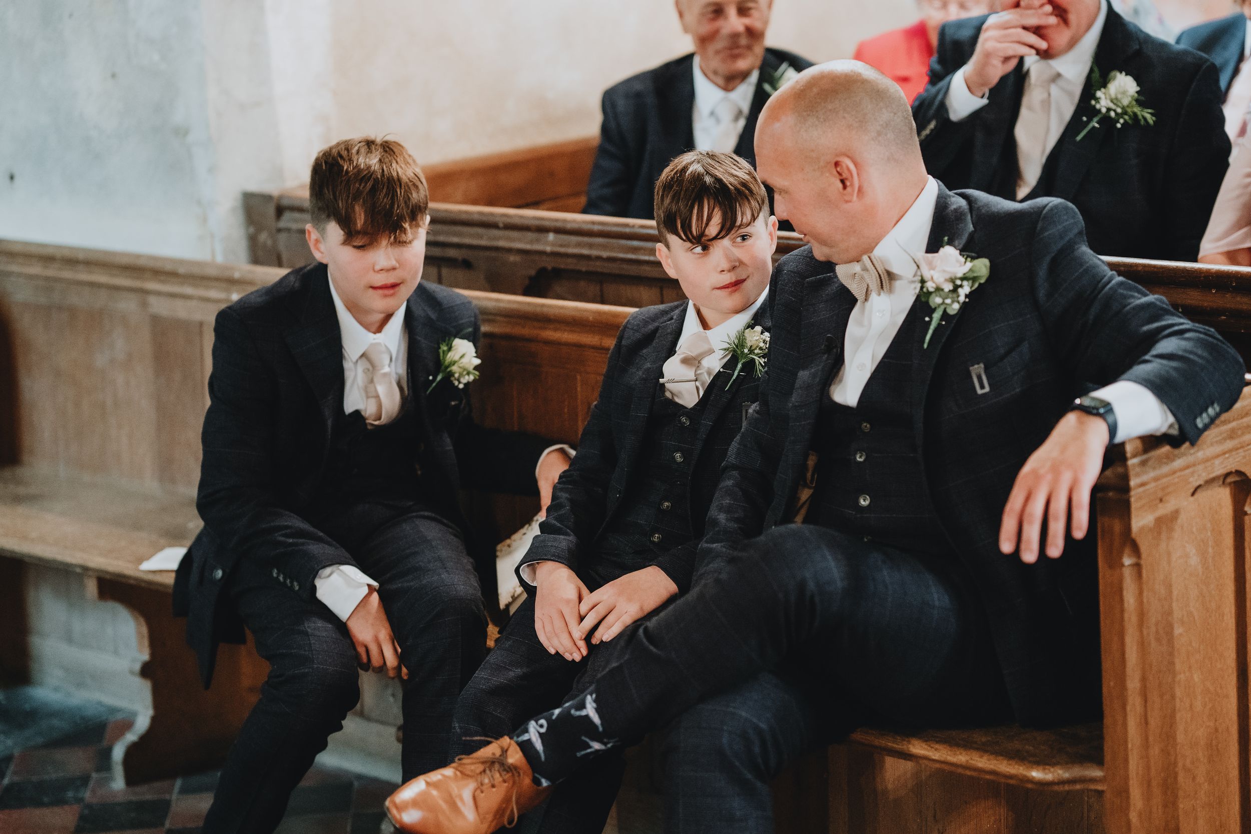 Three people in formal black attire and bow ties sit together on a wooden pew in a church.