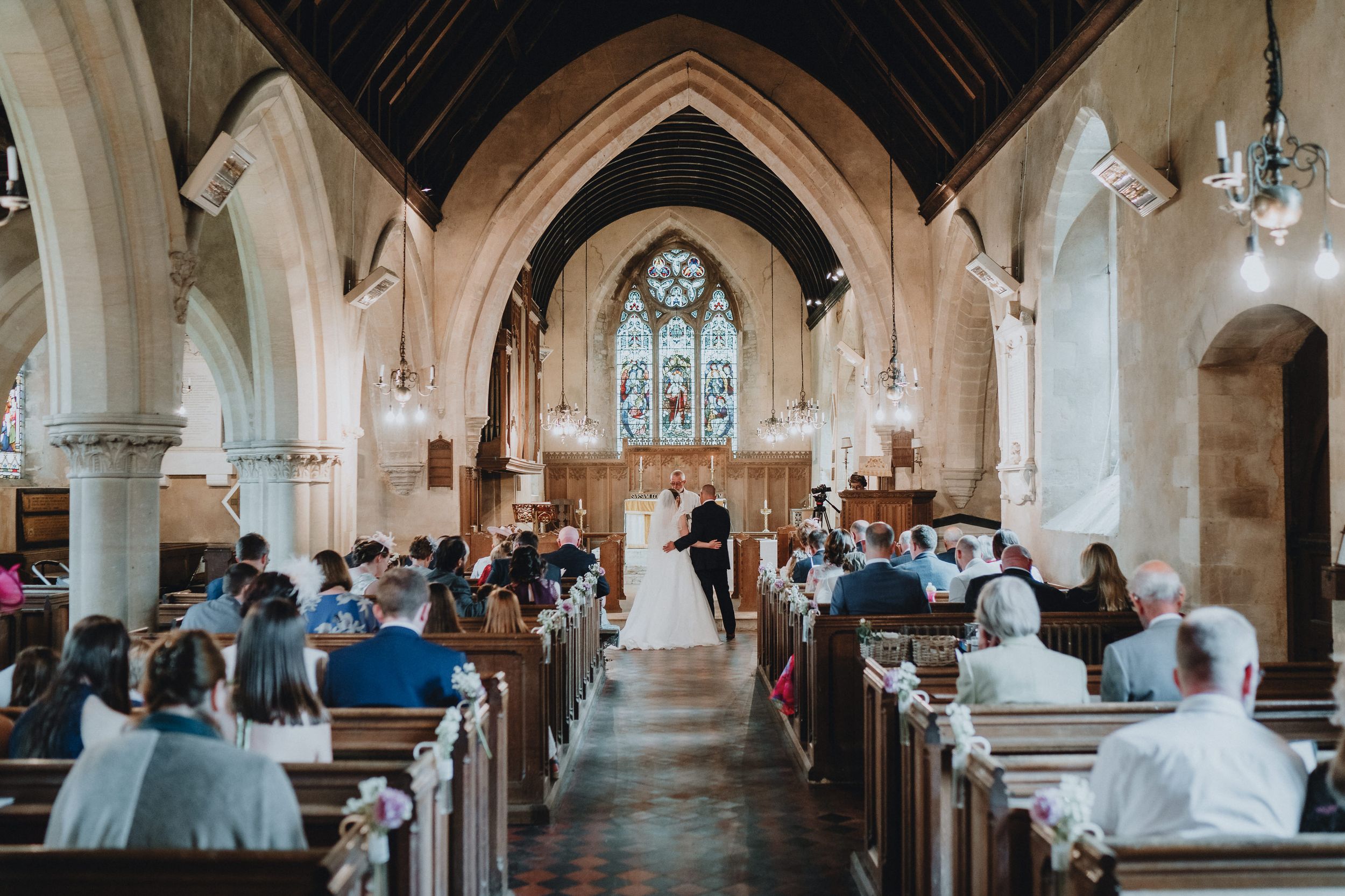 A traditional church wedding ceremony in progress with guests seated in wooden pews beneath gothic arches and stained glass.