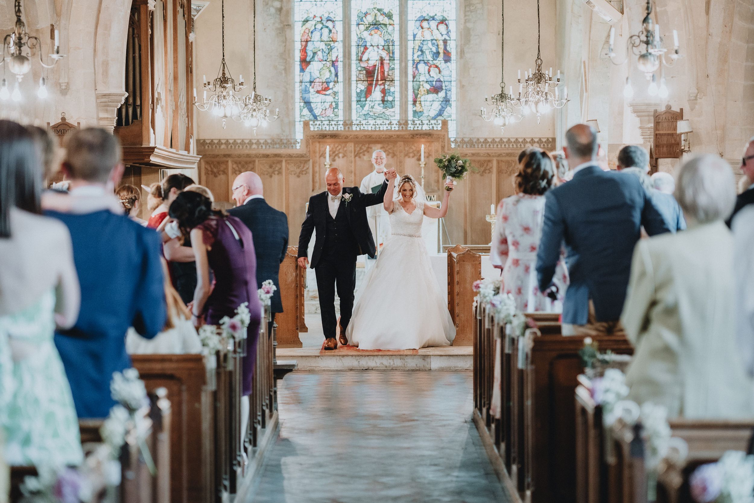 A wedding ceremony takes place in a historic stone church with stained glass windows and flower-lined wooden pews.
