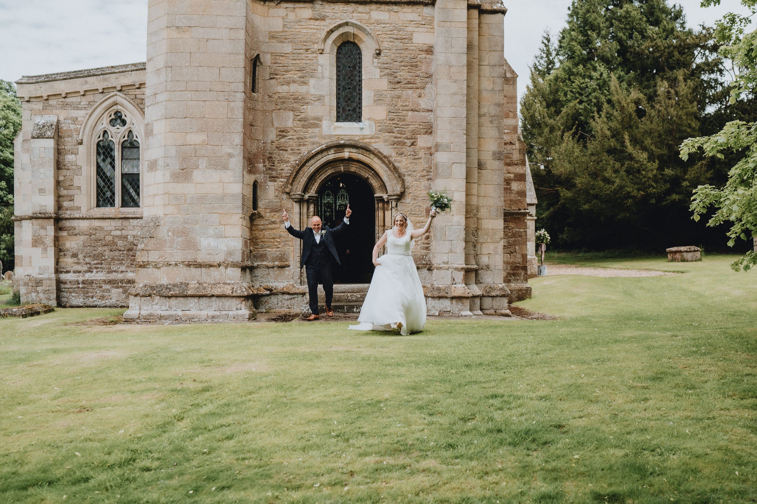 Two people in wedding attire stand outside the entrance of a historic stone church on a sunny day.