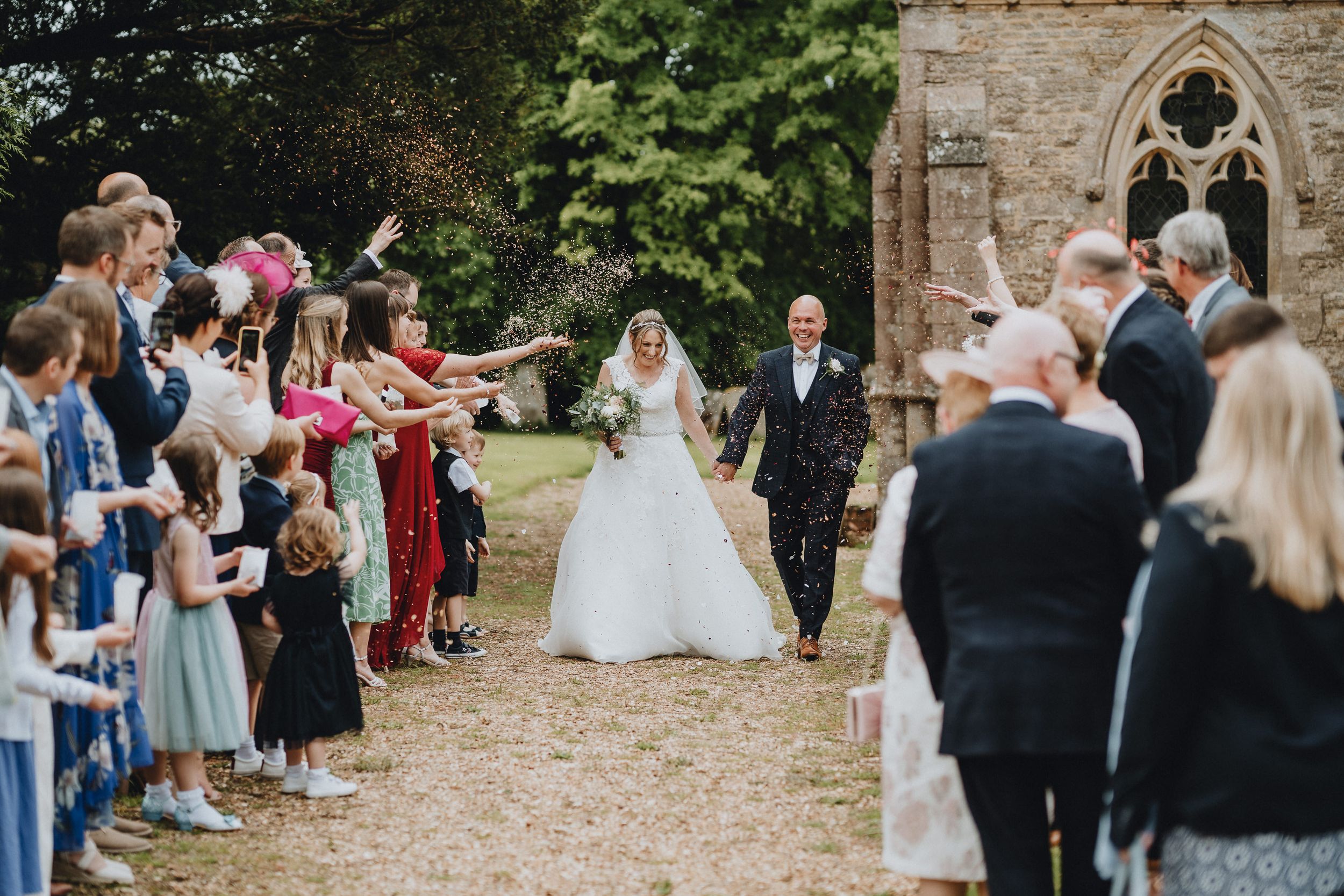 Wedding guests line a garden path outside a stone church as couple exits during a joyful outdoor celebration.