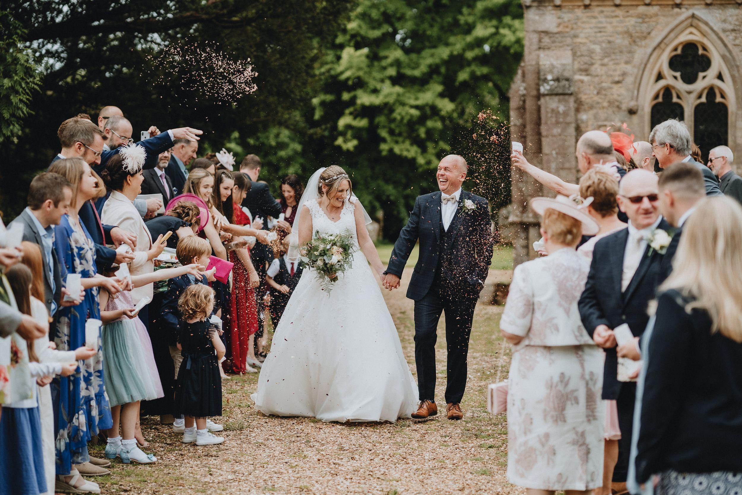Wedding guests shower newlyweds with confetti as they exit stone church along tree-lined path.