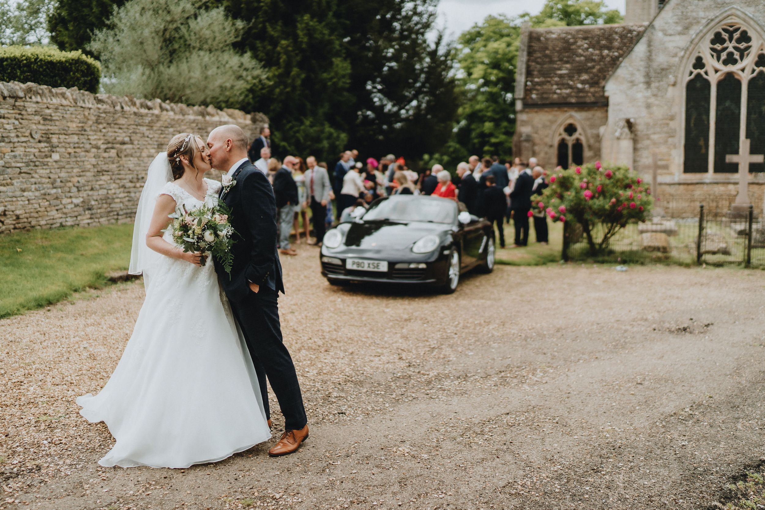 A classic black Porsche sports car parked outside a stone church during a wedding ceremony.