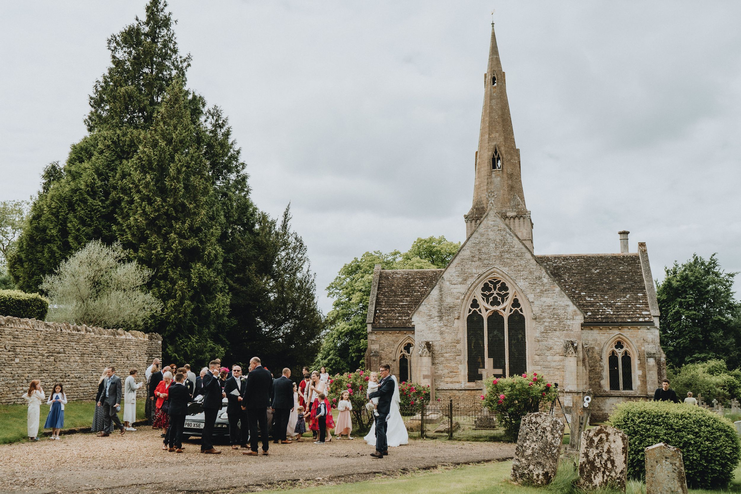Small stone church with spire hosts outdoor wedding ceremony gathering in countryside setting with overcast sky.
