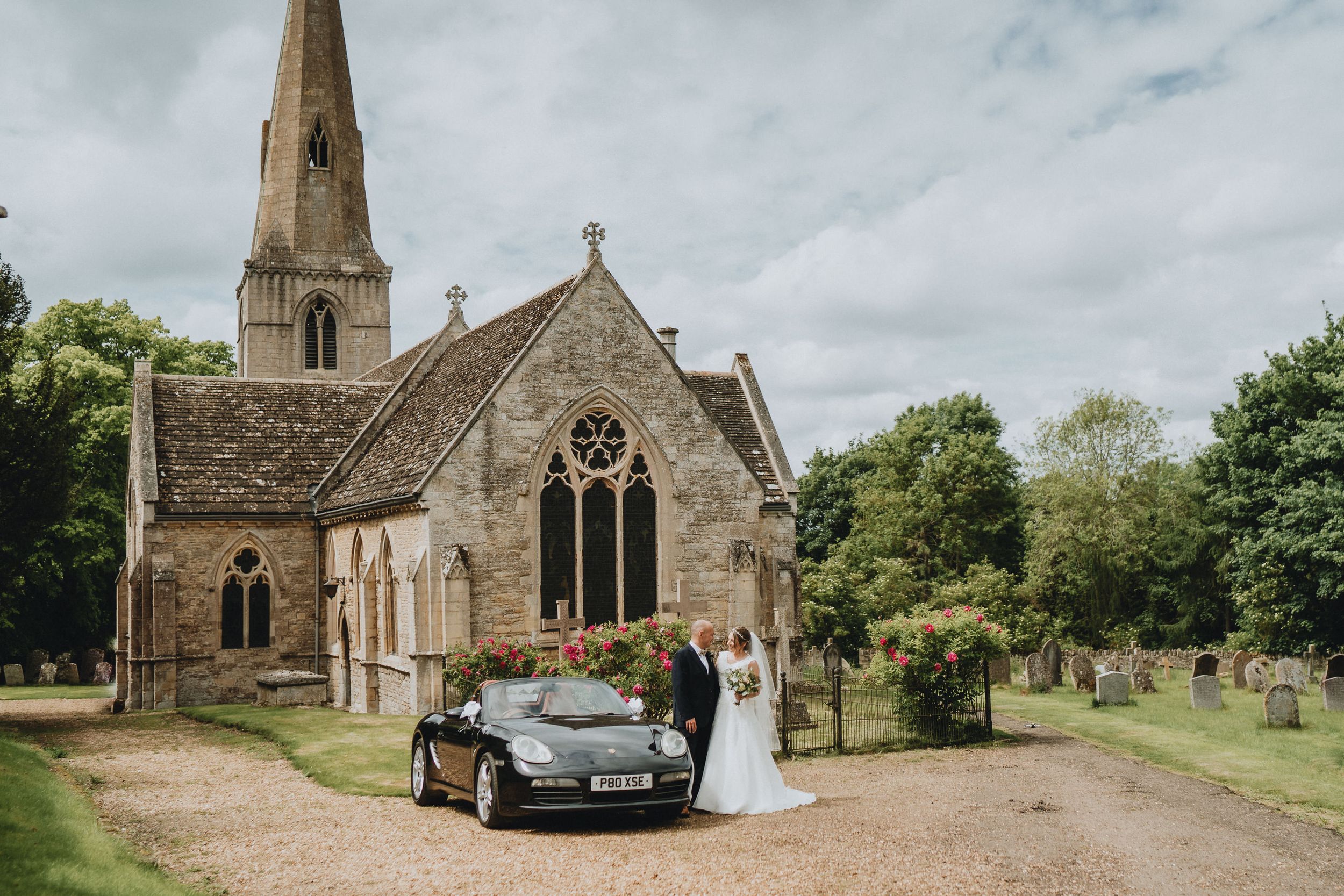 Classic stone church with tall spire and vintage car parked outside during wedding ceremony.