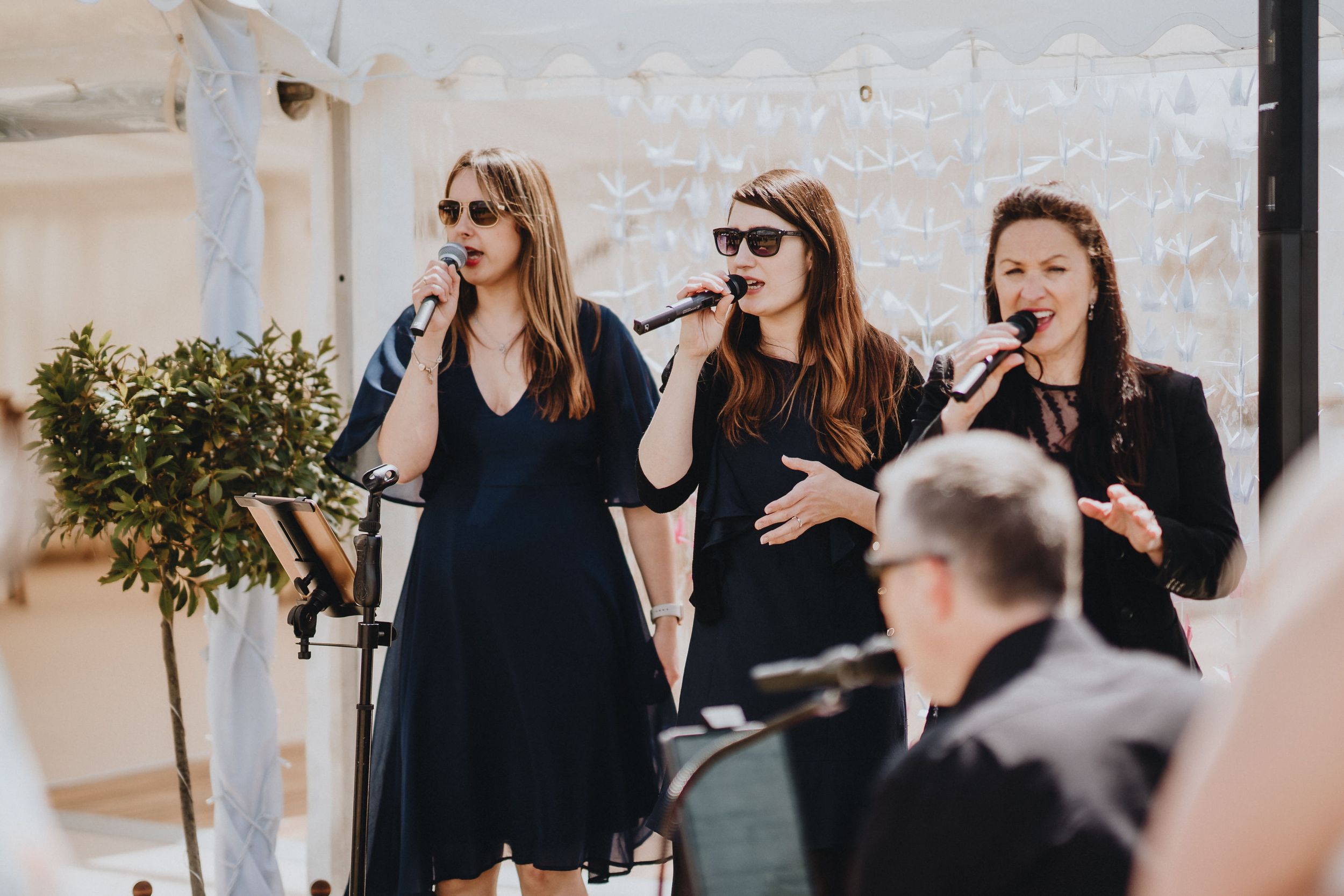Three performers in black dresses singing together at an event while accompanied by musicians.