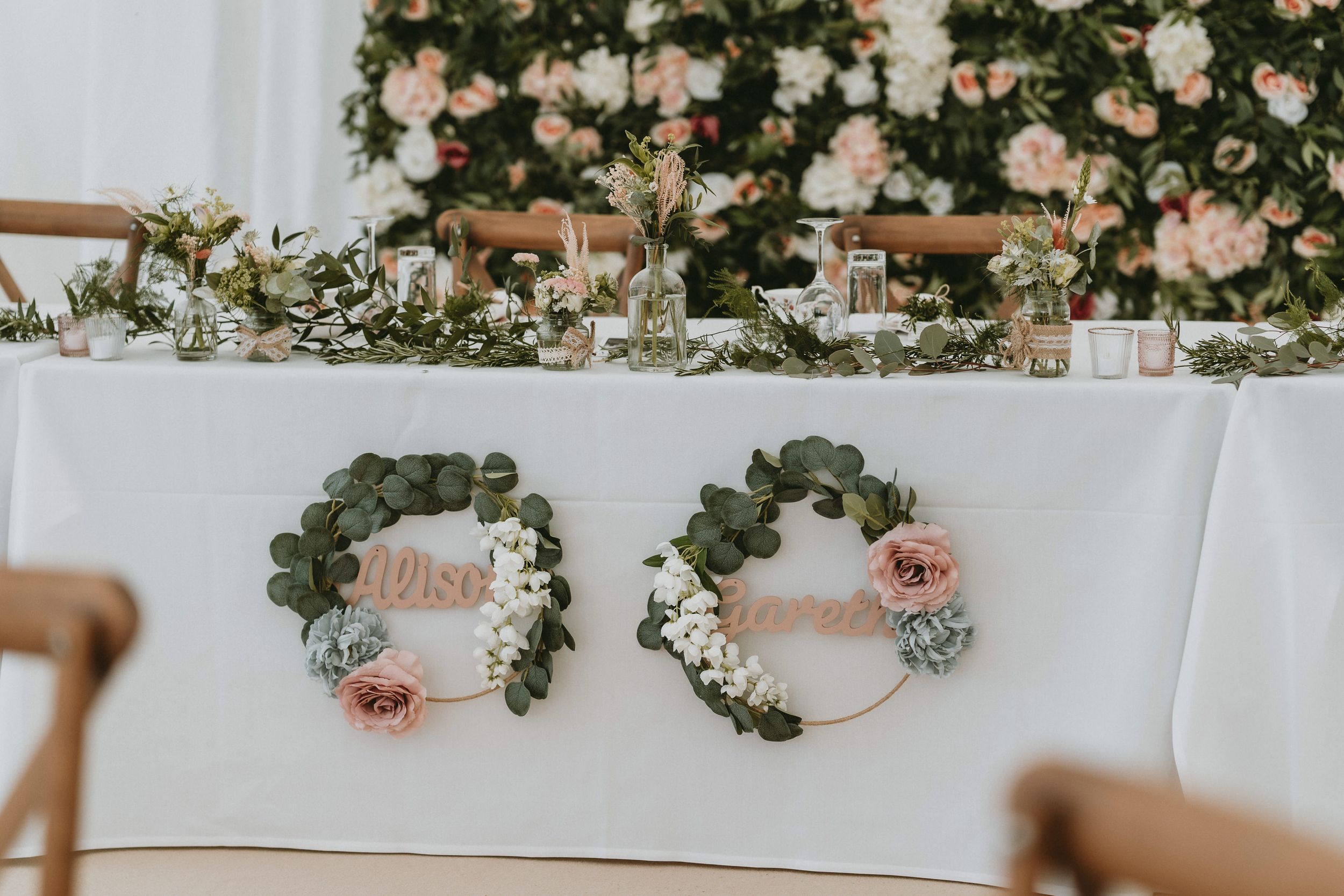 Wedding table decor with floral wreaths reading 'Mrs' adorned with pink roses against white tablecloth and greenery backdrop.