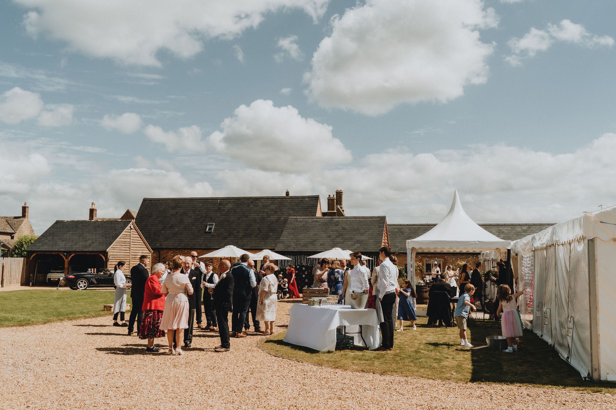 Outdoor market event with white vendor tents and crowd gathered on gravel path near rustic stone buildings under blue sky.