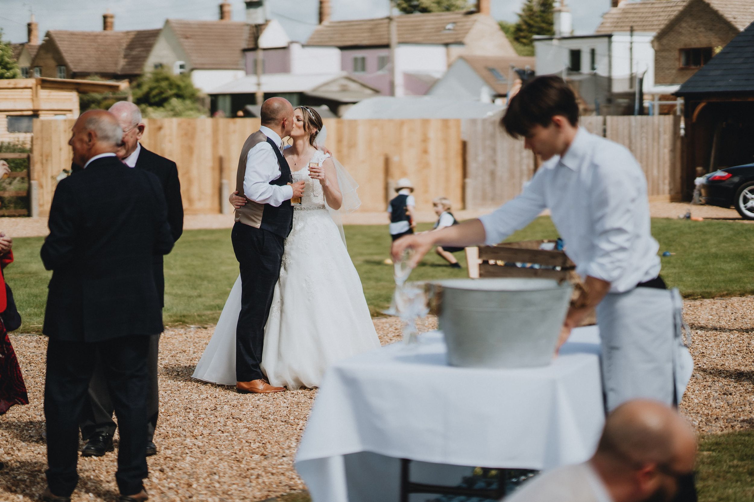 A casual outdoor wedding reception with guests gathering around while a staff member tends to a beverage station.