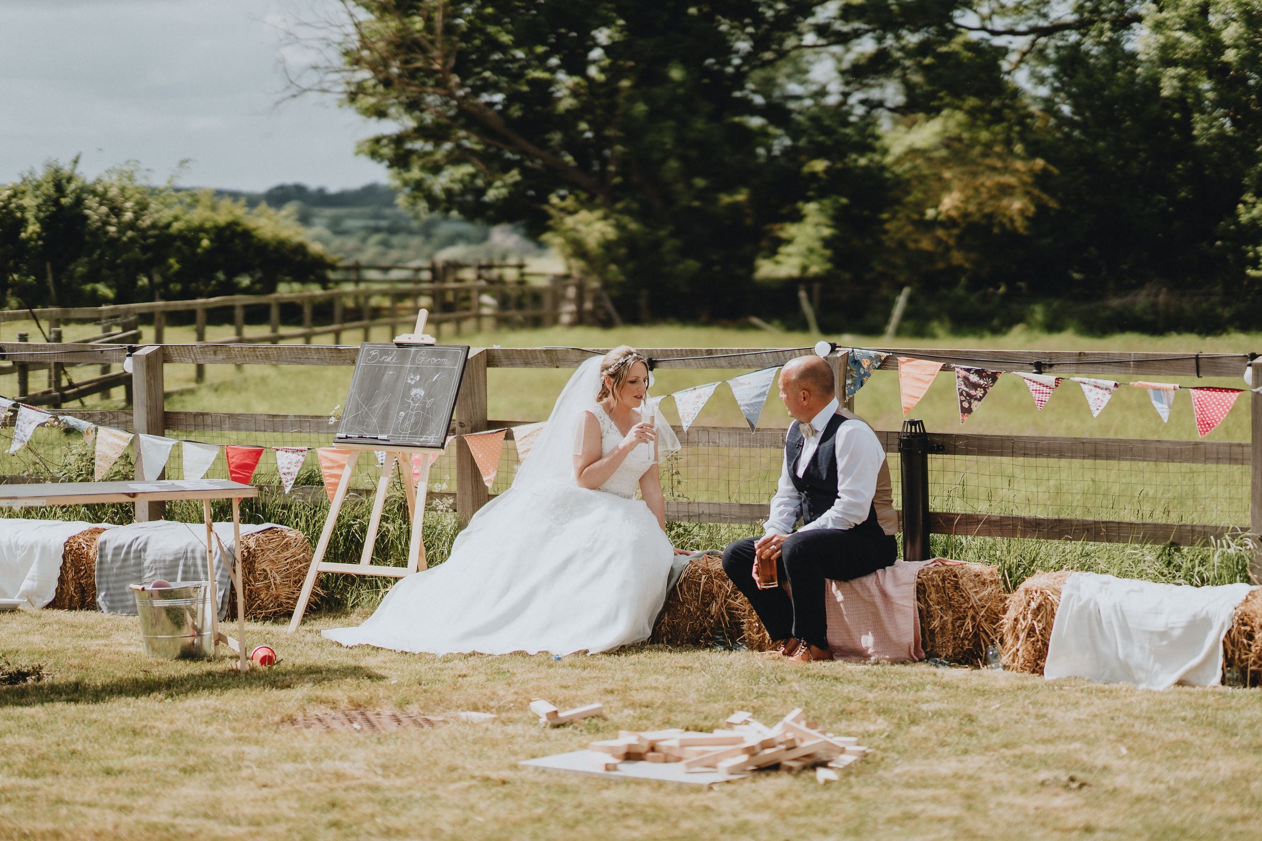 A rustic outdoor wedding scene with hay bales, wooden fencing, and pastel bunting flags decorating a sunny countryside setting.