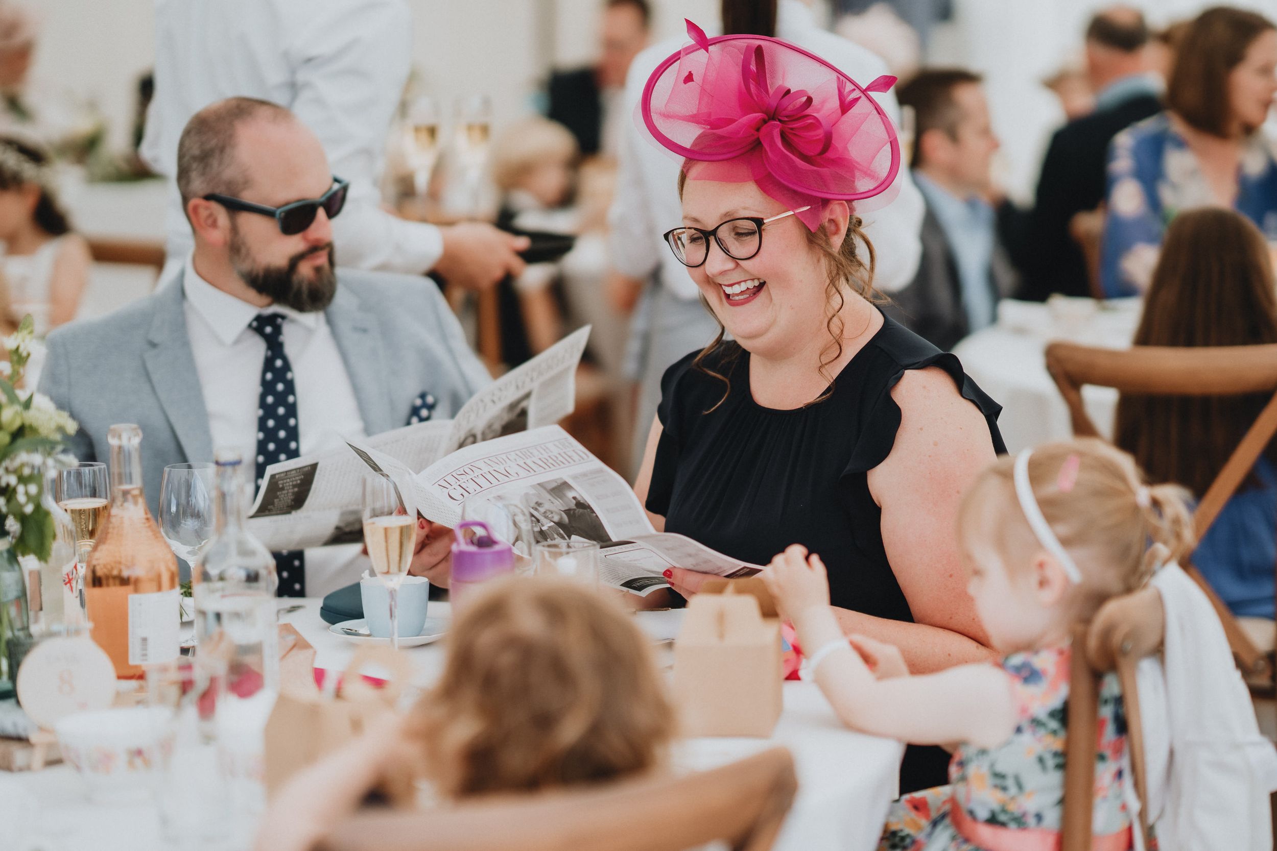 Wedding guests enjoy conversation at reception table, with woman in black dress wearing bright pink fascinator.