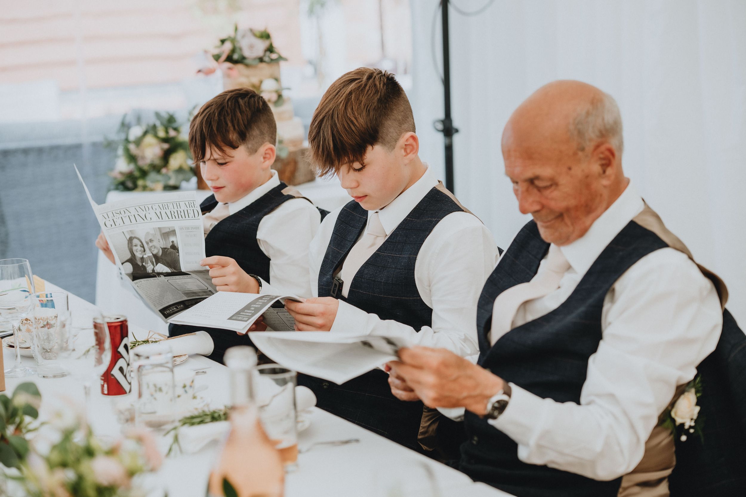 Three people in matching formal vests read their papers while seated at a decorated dining table.