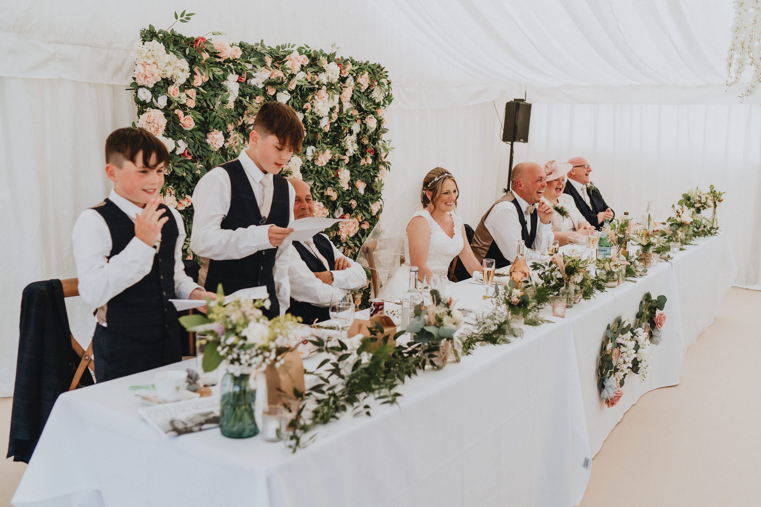 Wedding party sits together at a decorated head table adorned with floral arrangements in a white reception tent.