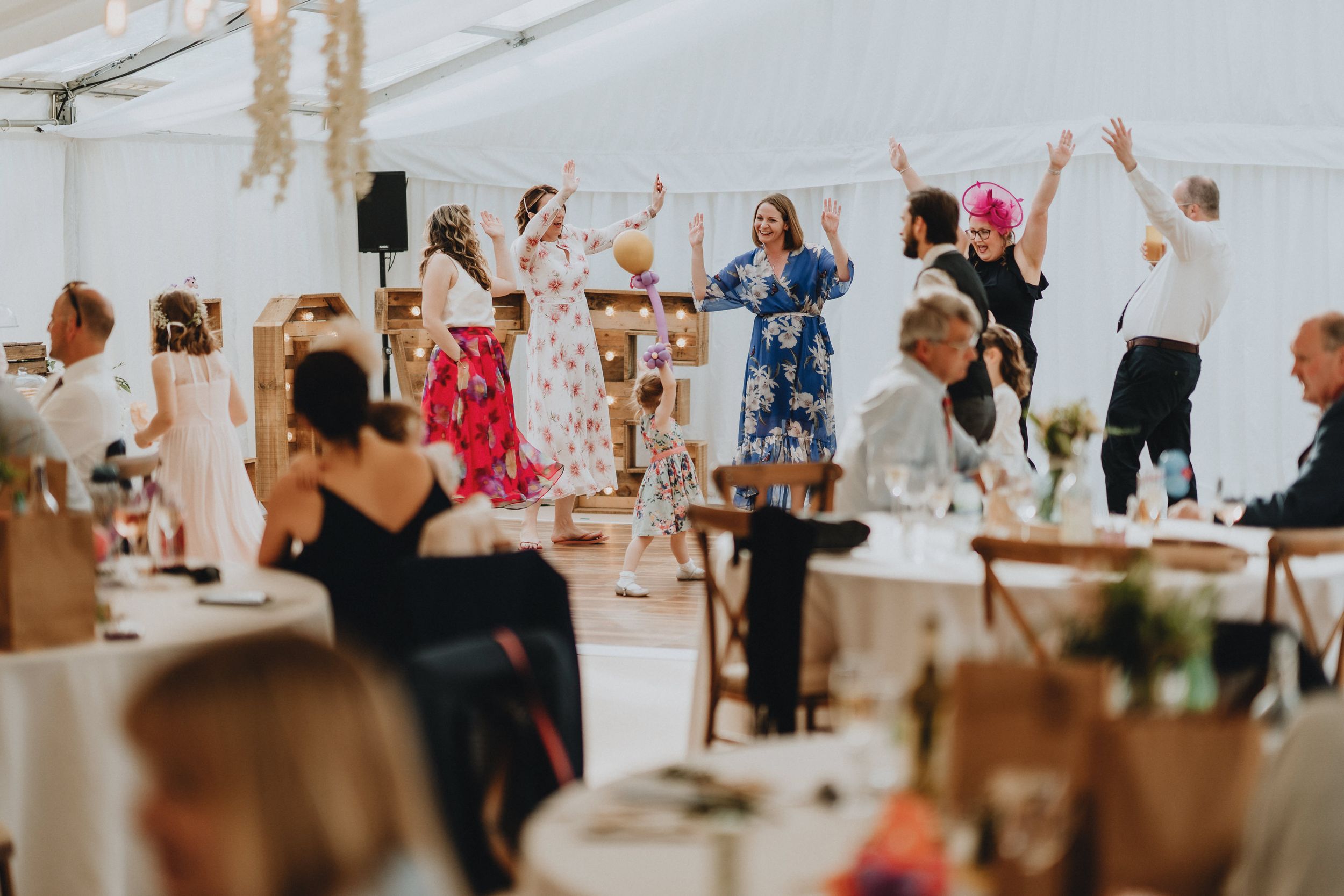 Party guests dancing enthusiastically together under a white tent at an outdoor reception event.