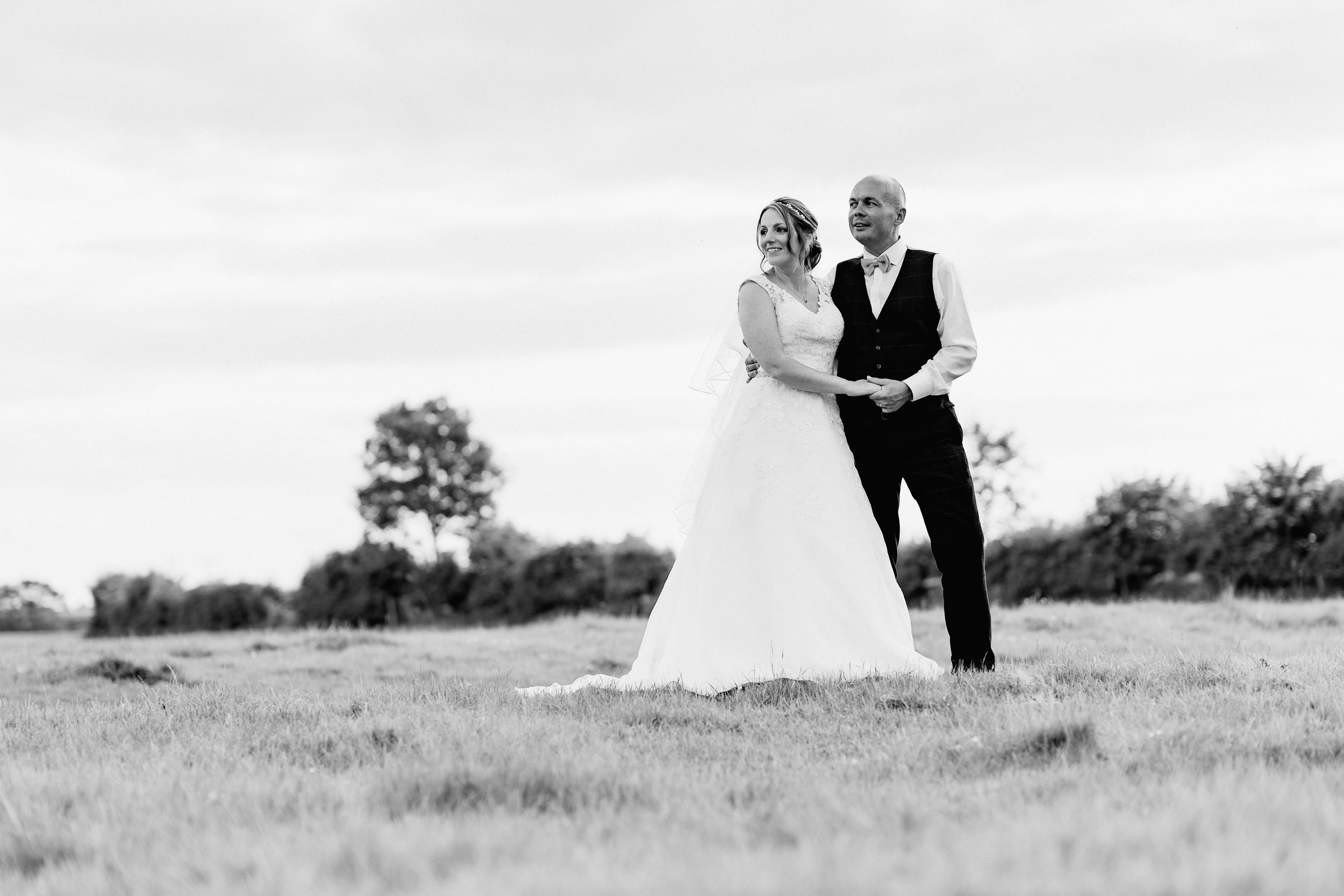 A couple stands together on a hilltop meadow in black and white.