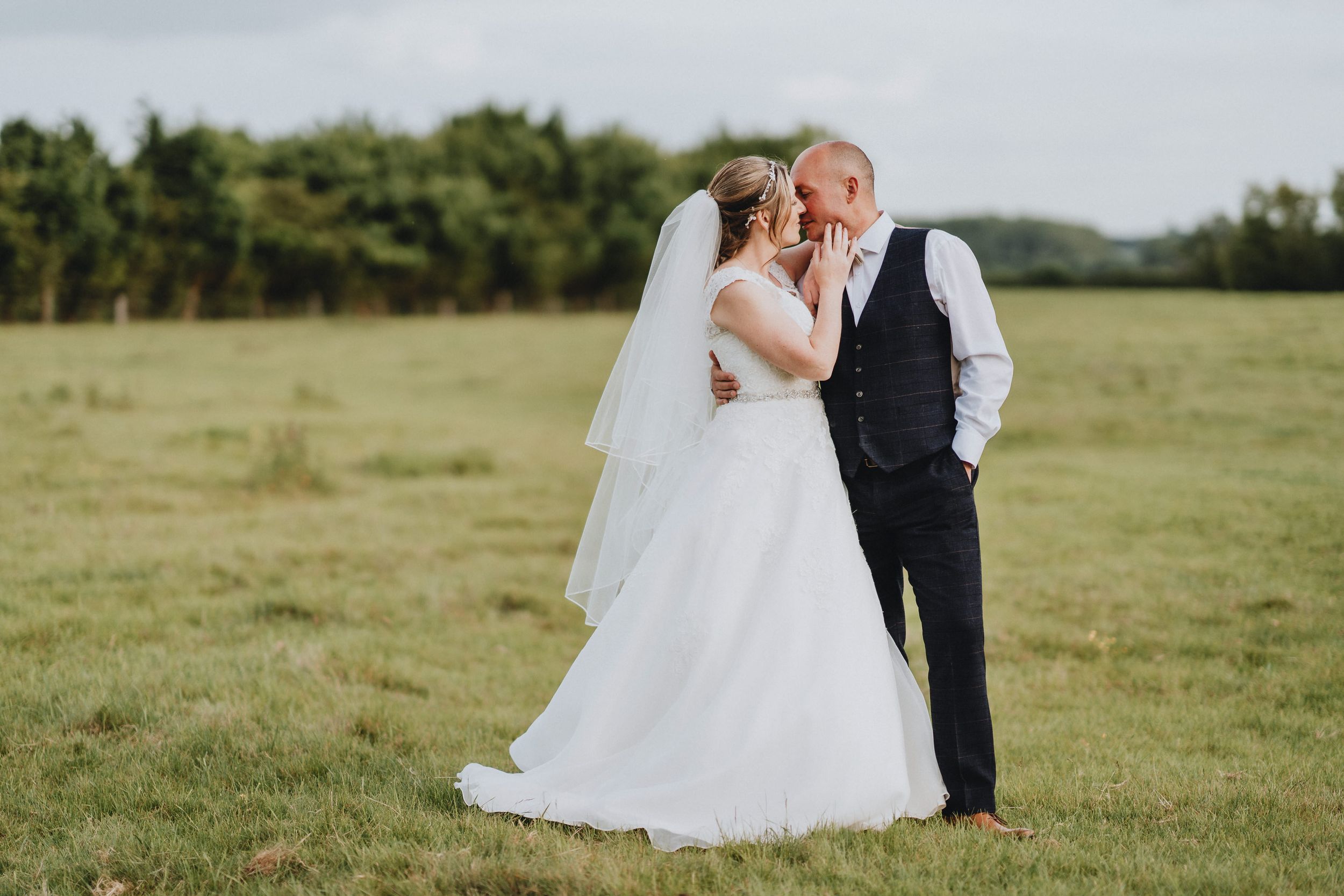 A romantic wedding photo captured in a green field with natural lighting and a flowing white bridal gown.