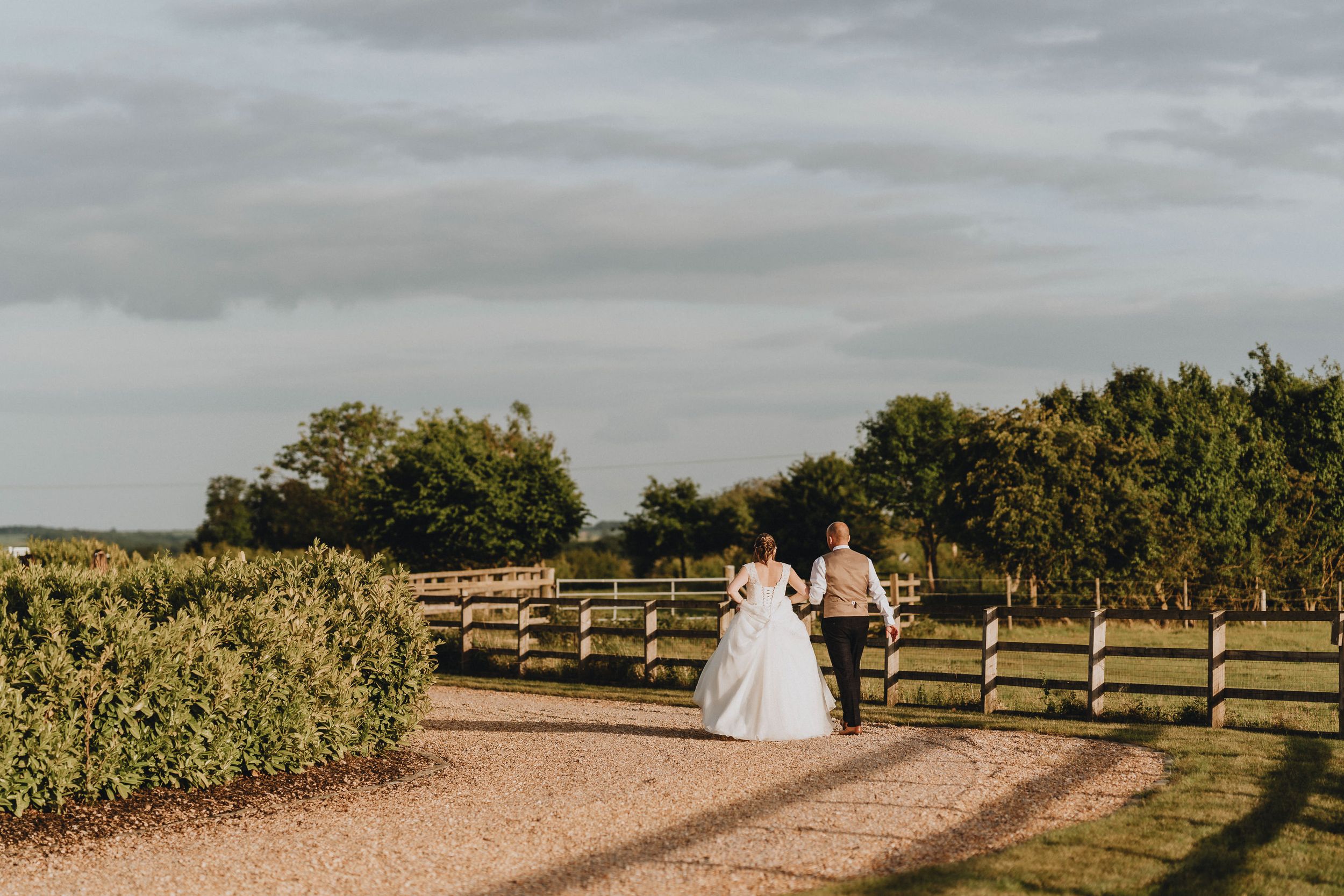A couple shares a romantic moment along a dirt path bordered by wooden fences and greenery at sunset.