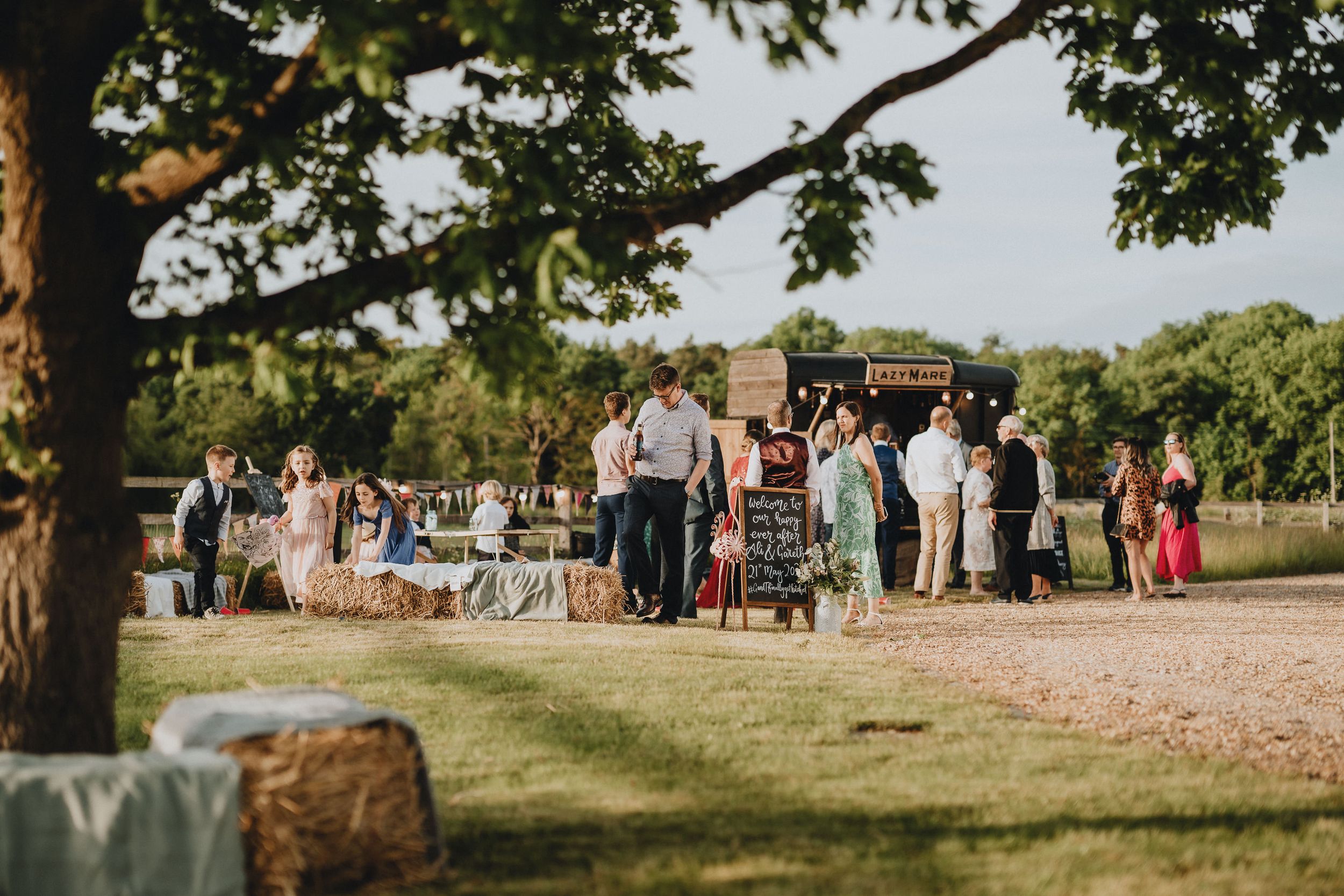 Wedding guests gather outdoors on a sunny day along a dirt path bordered by green trees and grass.