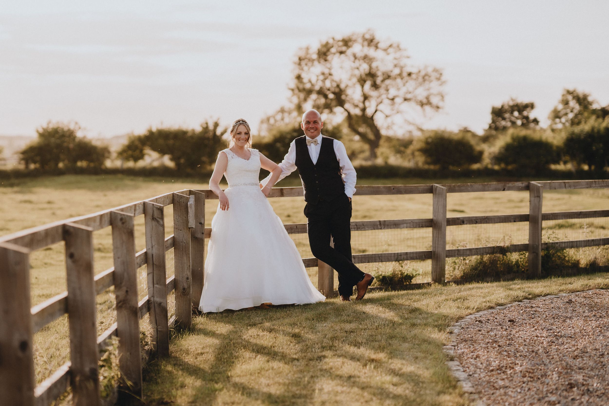 A couple in wedding attire holds hands while walking along a wooden fence at sunset on a rustic farm property.