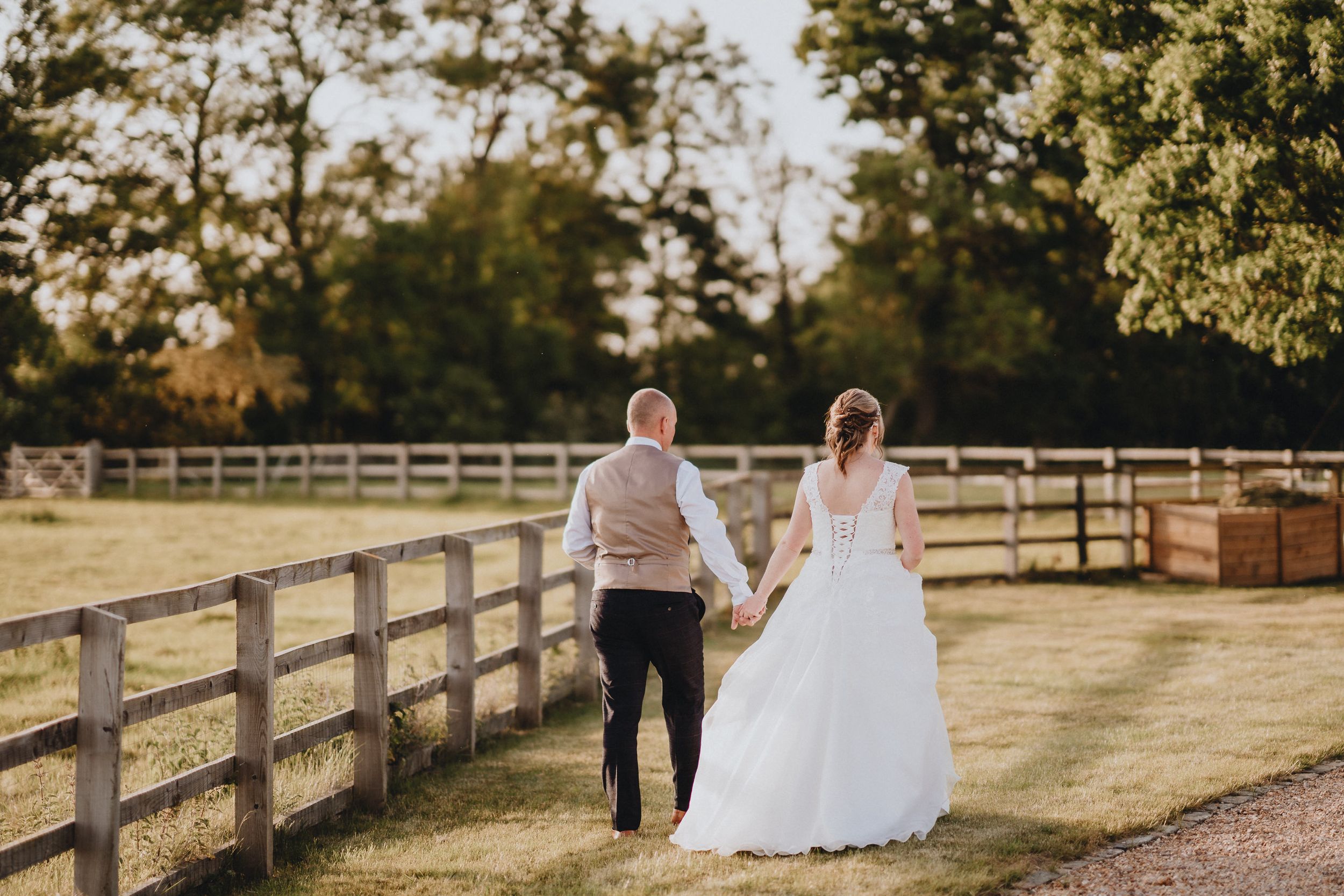 A couple holding hands walks along a wooden fence in a golden sunset at a rustic countryside venue.