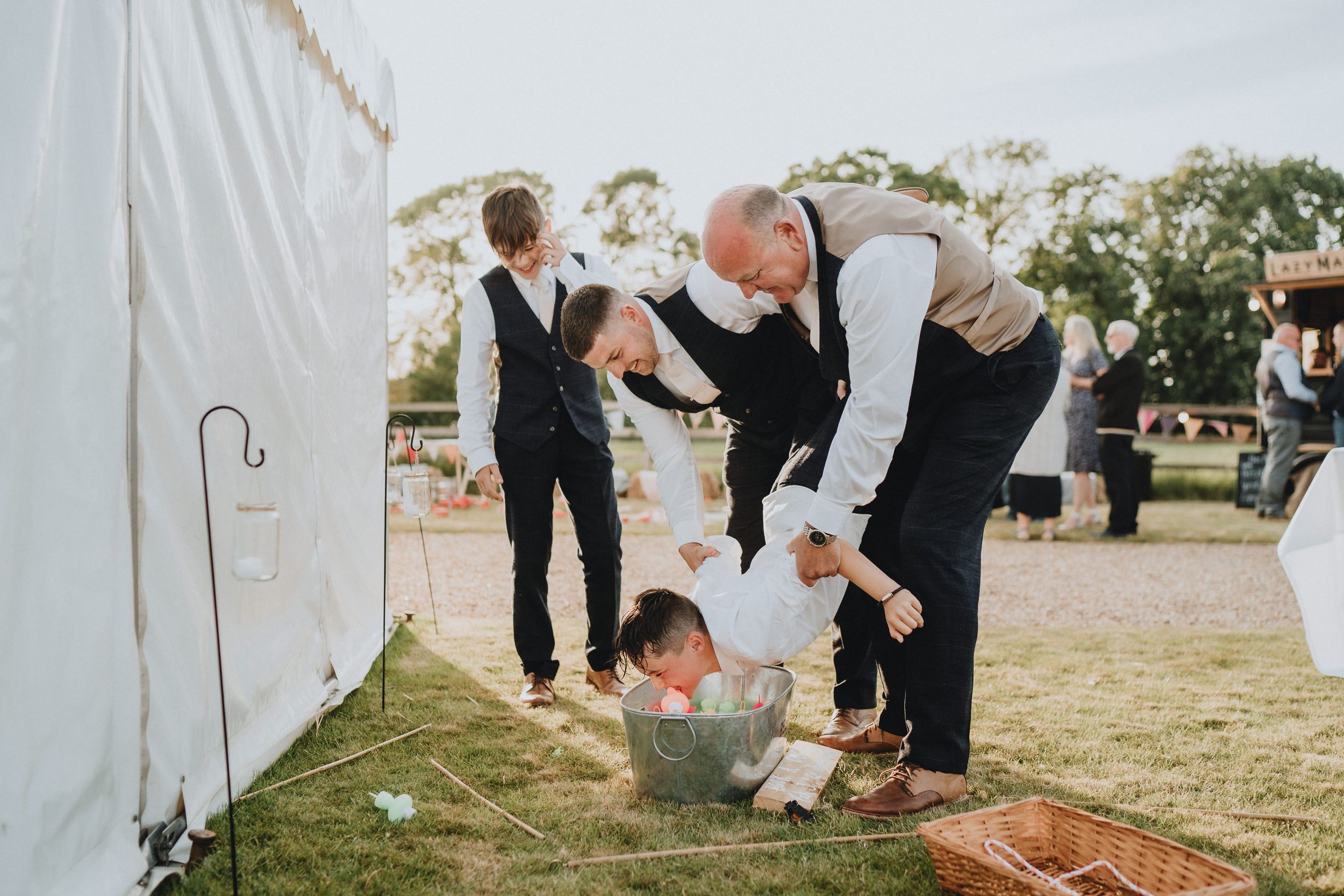 Wedding guests help a small child peeking under a white tent at an outdoor reception.