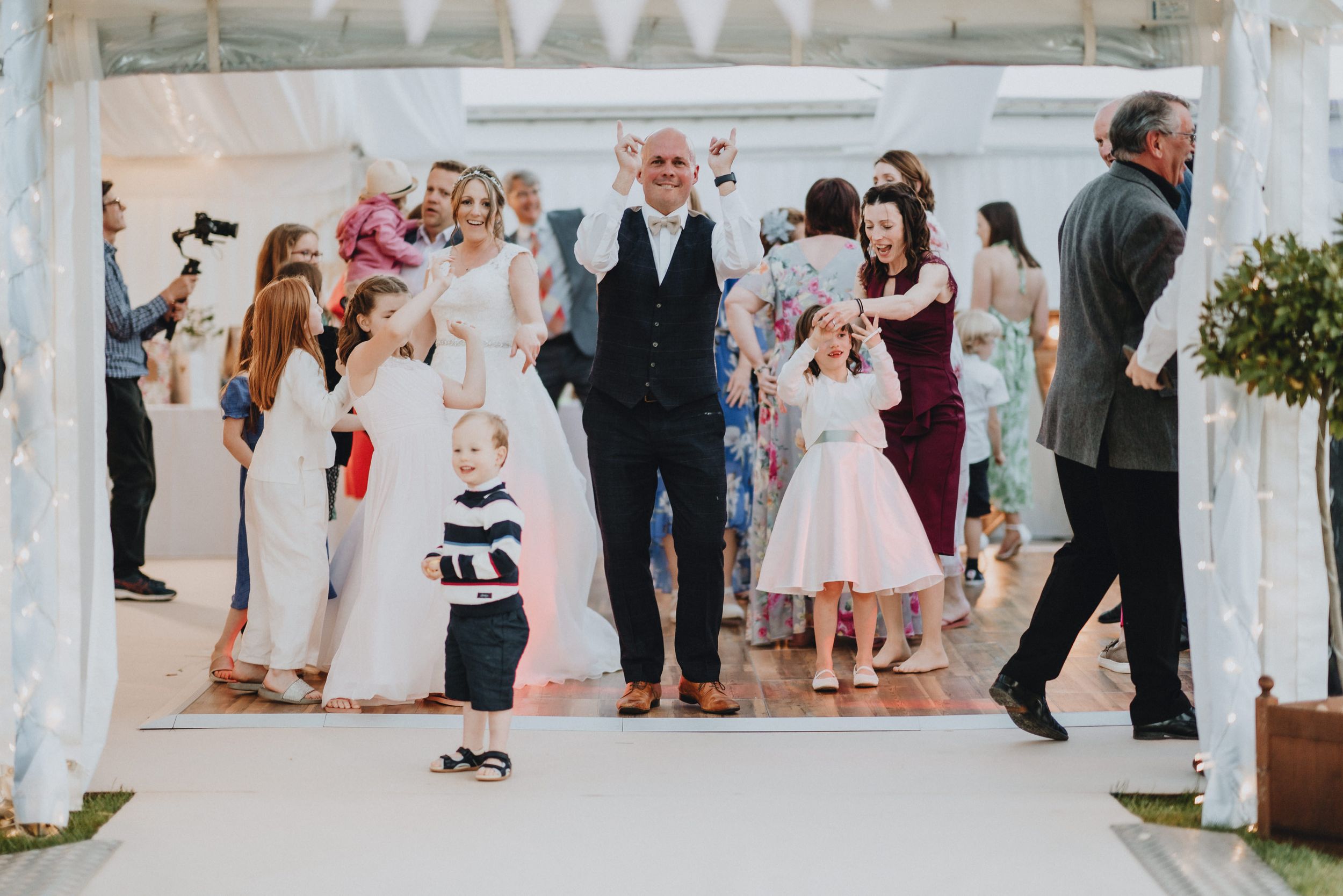 Guests gather under a white-draped outdoor pavilion during a wedding celebration on a bright sunny day.