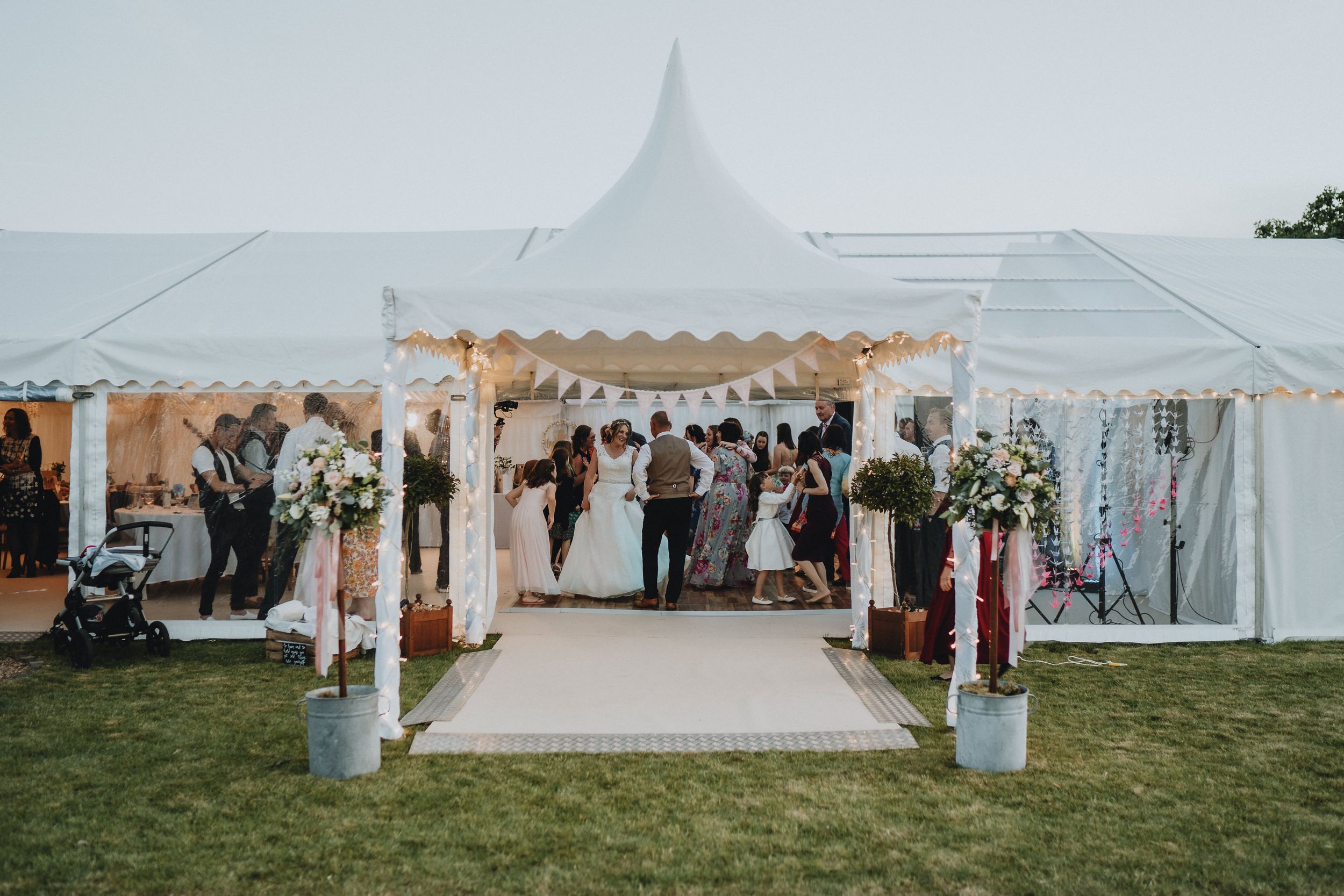 Wedding celebration under an elegant white tent with floral decorations and a raised platform on a grassy lawn.